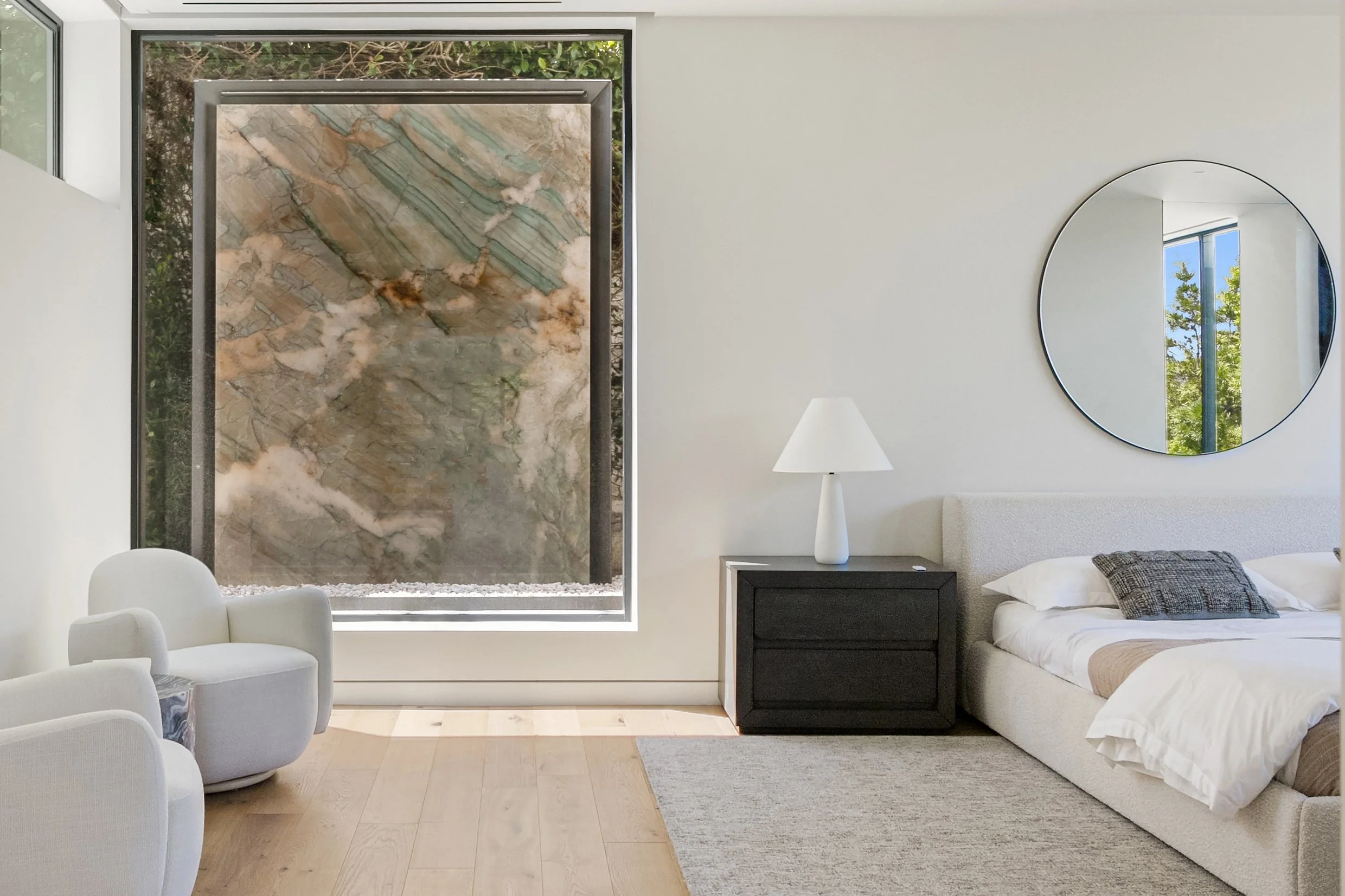 A modern, minimalist bedroom features a large, framed window showcasing a striking stone backdrop with green and brown veining. To the left, two stylish, round armchairs are positioned next to a small side table with a white lamp. A black nightstand