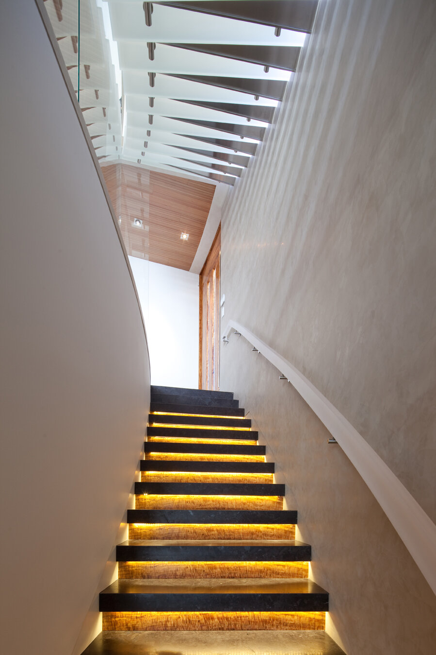 A modern staircase with dark steps featuring warm, glowing LED strip lights. The staircase has a sleek design, curving along a smooth wall with a handrail attached. Natural light streams in from a nearby window, highlighting the wood paneling on the