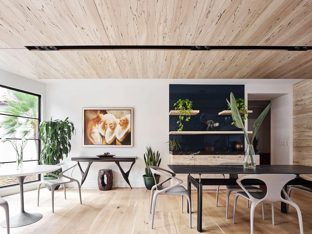 A modern dining area features a light wooden floor and a ceiling with distinctive wood grain patterns. On one side, large windows let in natural light, showcasing a tropical plant outside. A colorful artwork depicting three individuals with elaborate