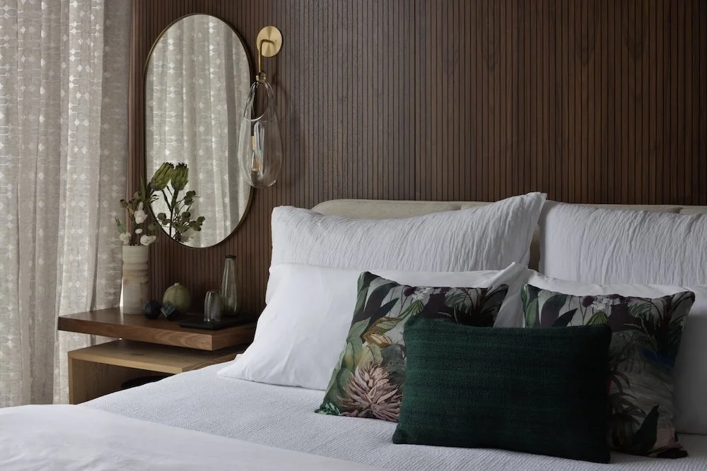 A cozy bedroom featuring a wooden accent wall behind a bed with two large white pillows and three decorative pillows, including one with a floral design and a dark green textured one. To the left, a round mirror reflects the light filtered through sh