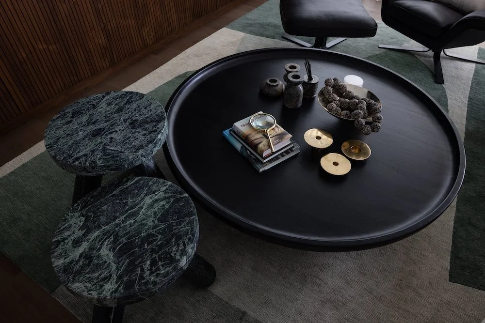 A modern living space featuring a round black coffee table at the center, adorned with decorative objects, including a magnifying glass, several small vases, and bowls containing natural elements. Stacked books are placed on the table. Two marble-sty