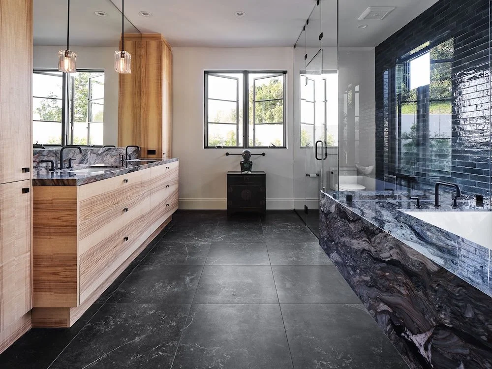 A modern bathroom featuring sleek design elements. On the left side, a wooden vanity with multiple drawers is topped with a dark patterned countertop and fitted with black faucets. Above, two glass pendant lights hang down. The wall on the right has