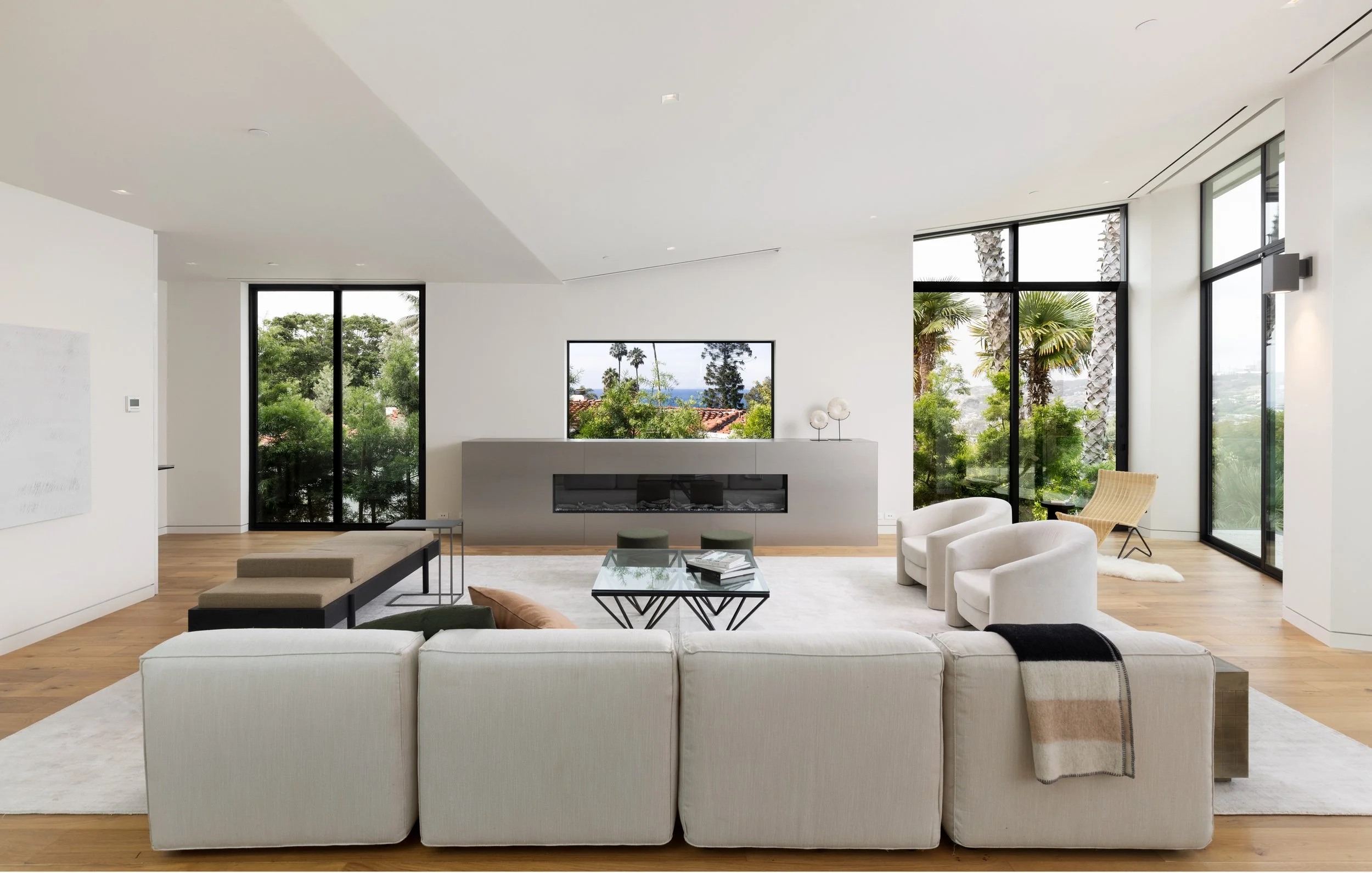 A modern, minimalist living room features a large, light-colored sectional sofa with a patterned throw blanket draped over the back. In the center, there's a glass coffee table with a few magazines on it. Surrounding the table are two white armchairs