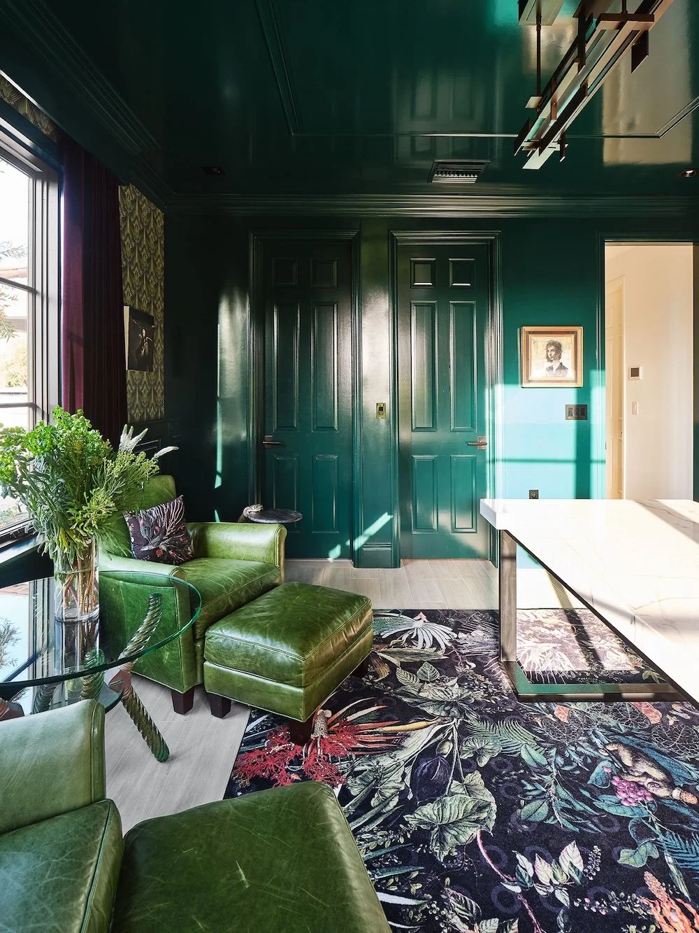A stylish interior room features deep green paneled walls and a similarly painted ceiling. Two closed double doors are prominent on one side, while a large window with red curtains lets in natural light. A large area rug with a colorful floral design