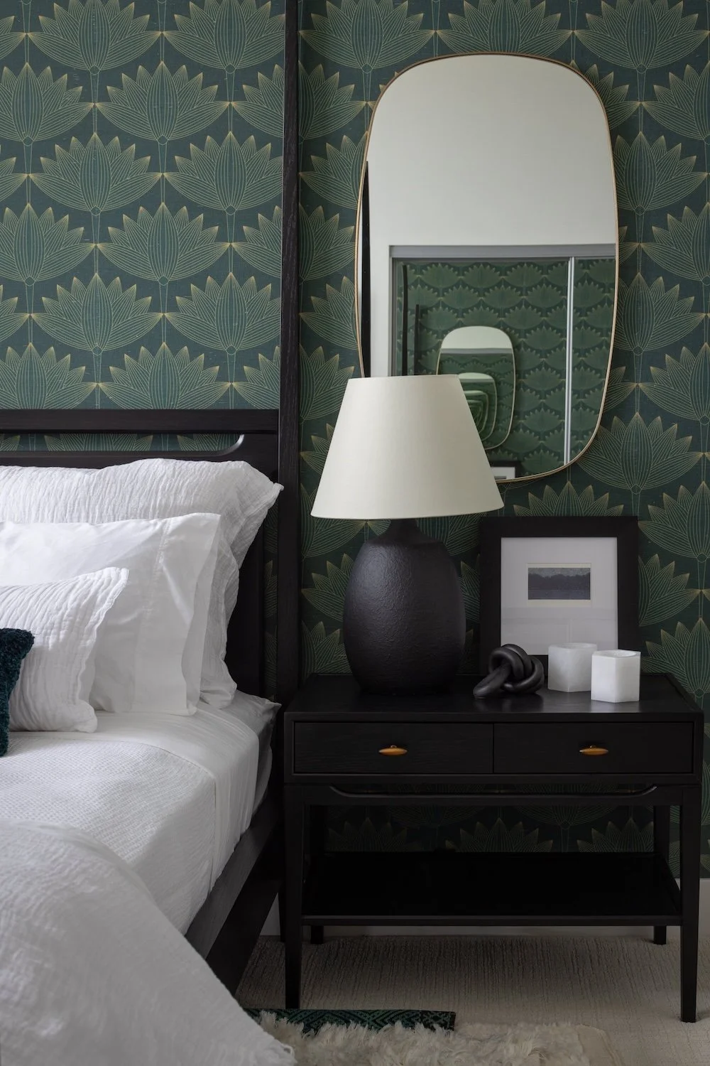 A modern bedroom scene featuring a black bed frame with white bedding, positioned against a dark green wall adorned with a floral pattern. On the right, a bedside table with two drawers holds a textured black lamp with a white shade, a framed picture