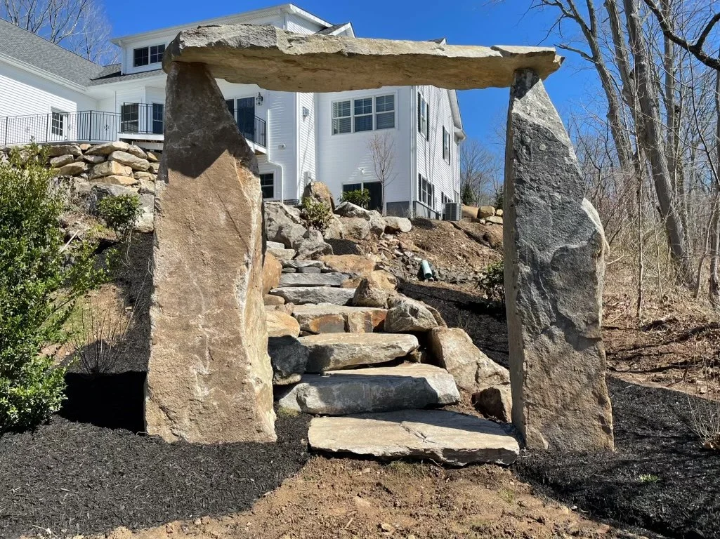 Rustic and primal charm are added to this entryway with granite stone steps framed at their entrance with a monolithic stone archway inspired by the beauty and balance of Stonehenge