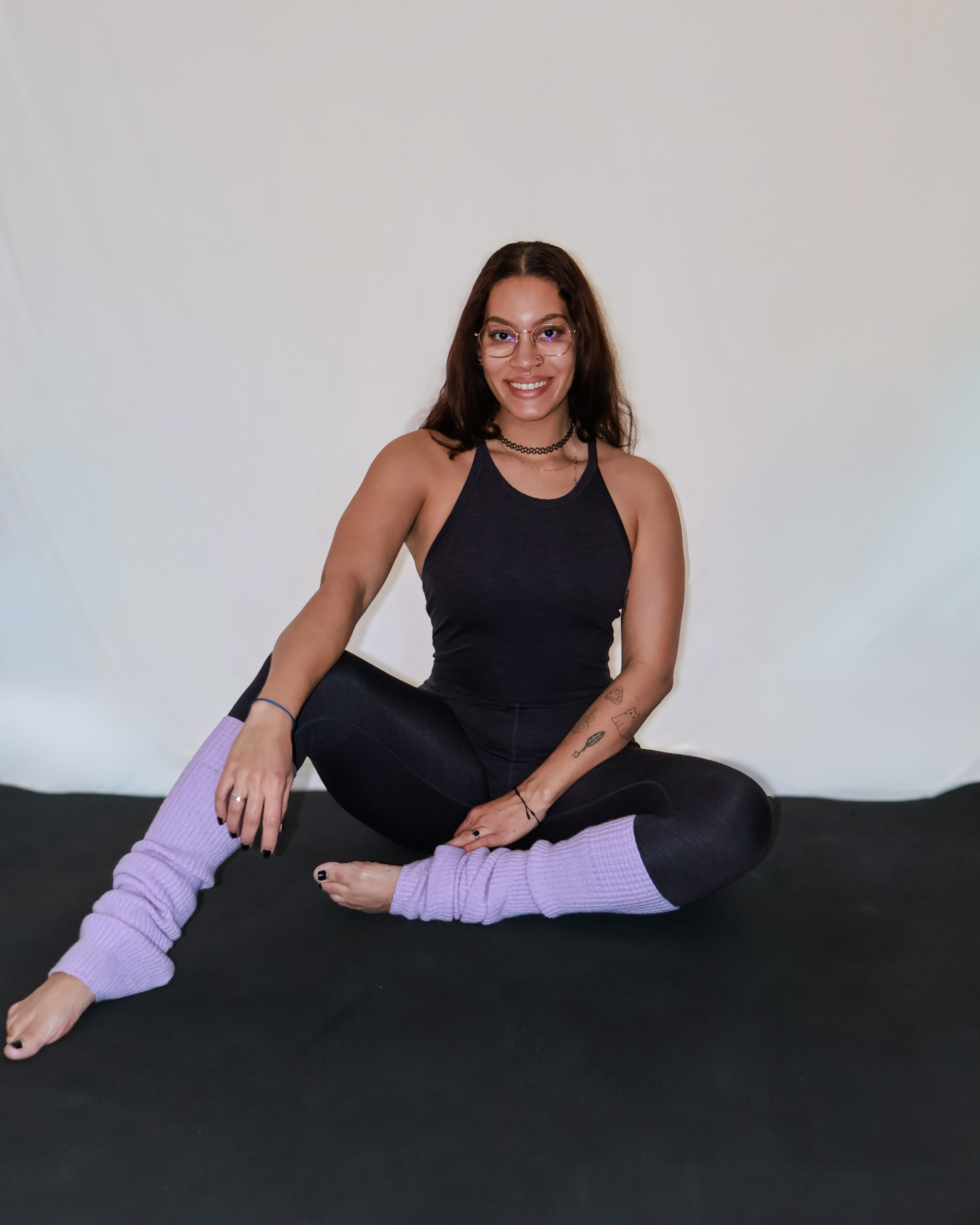 A young woman with glasses, tattoos, and dark hair sitting cross-legged on a black surface, smiling, wearing a black tank top, black leggings, and lavender leg warmers.