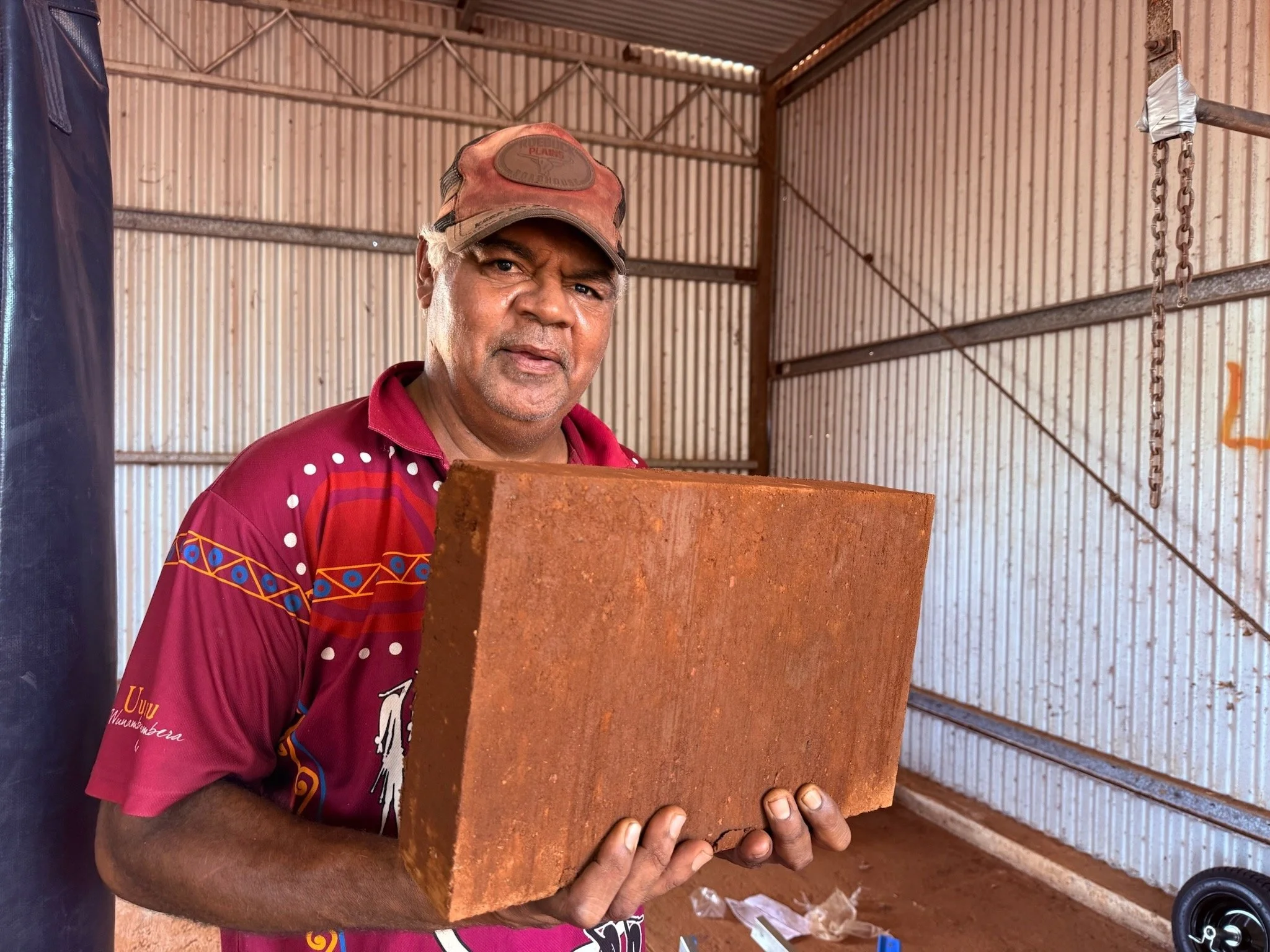 Aboriginal man stands in a metal shed holding a compressed earth block