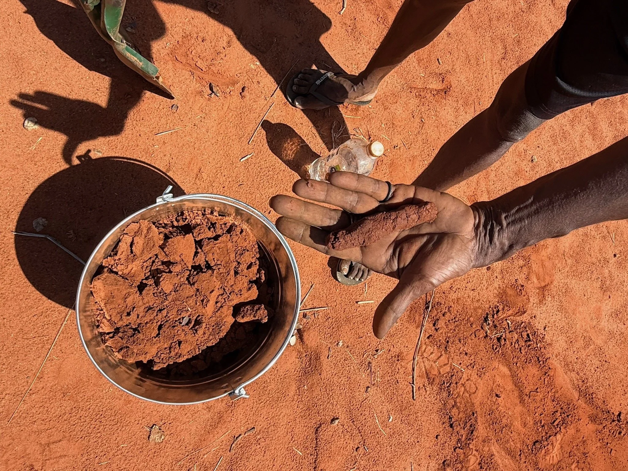 A metal bucket full of earth is on the ground and above it a brown-skinned hand holds a wad of wet earth to demonstrate the consistency is good for earth brick making