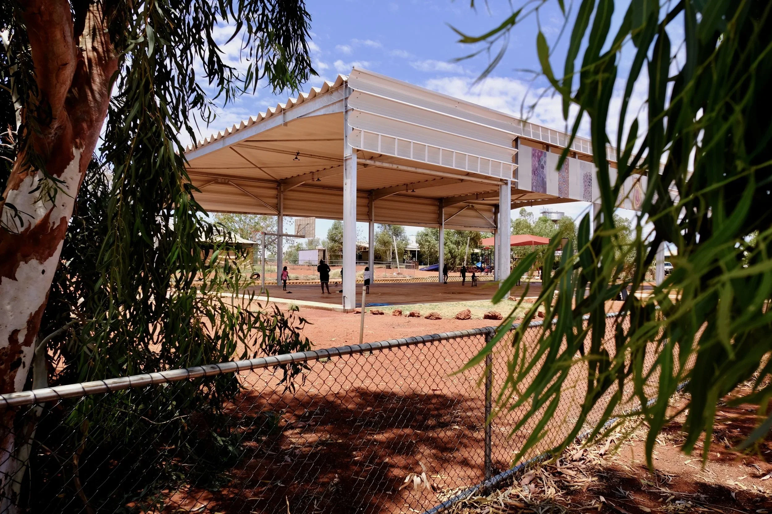 Through the trees a metal-roofed basketball court is visible. It has Aboriginal paintings along the vertical awning