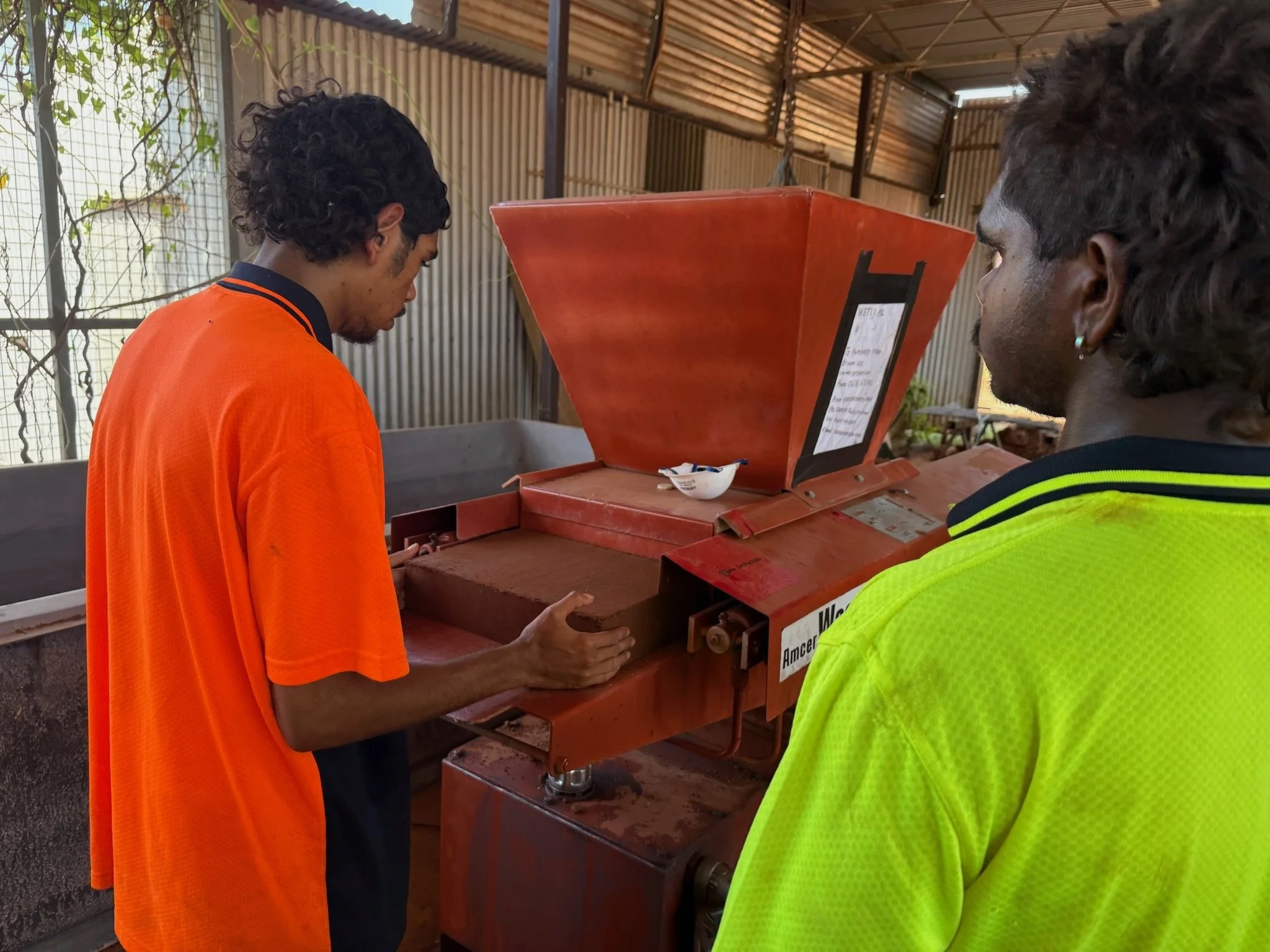 Two men stand in front of a machine for compressing earth into blocks. One is taking a brick as it emerges from the machine