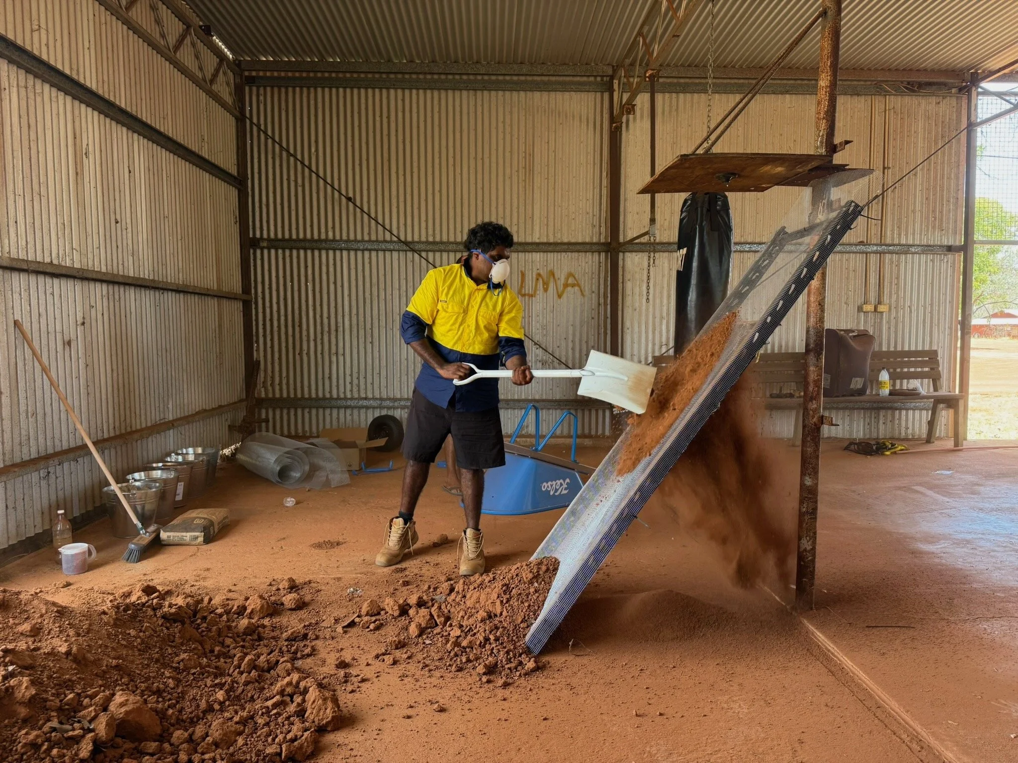 A man stands in front of a large sieve, using a spade to filter earth. He's wearing PPE and there are piles of earth and other tools around him