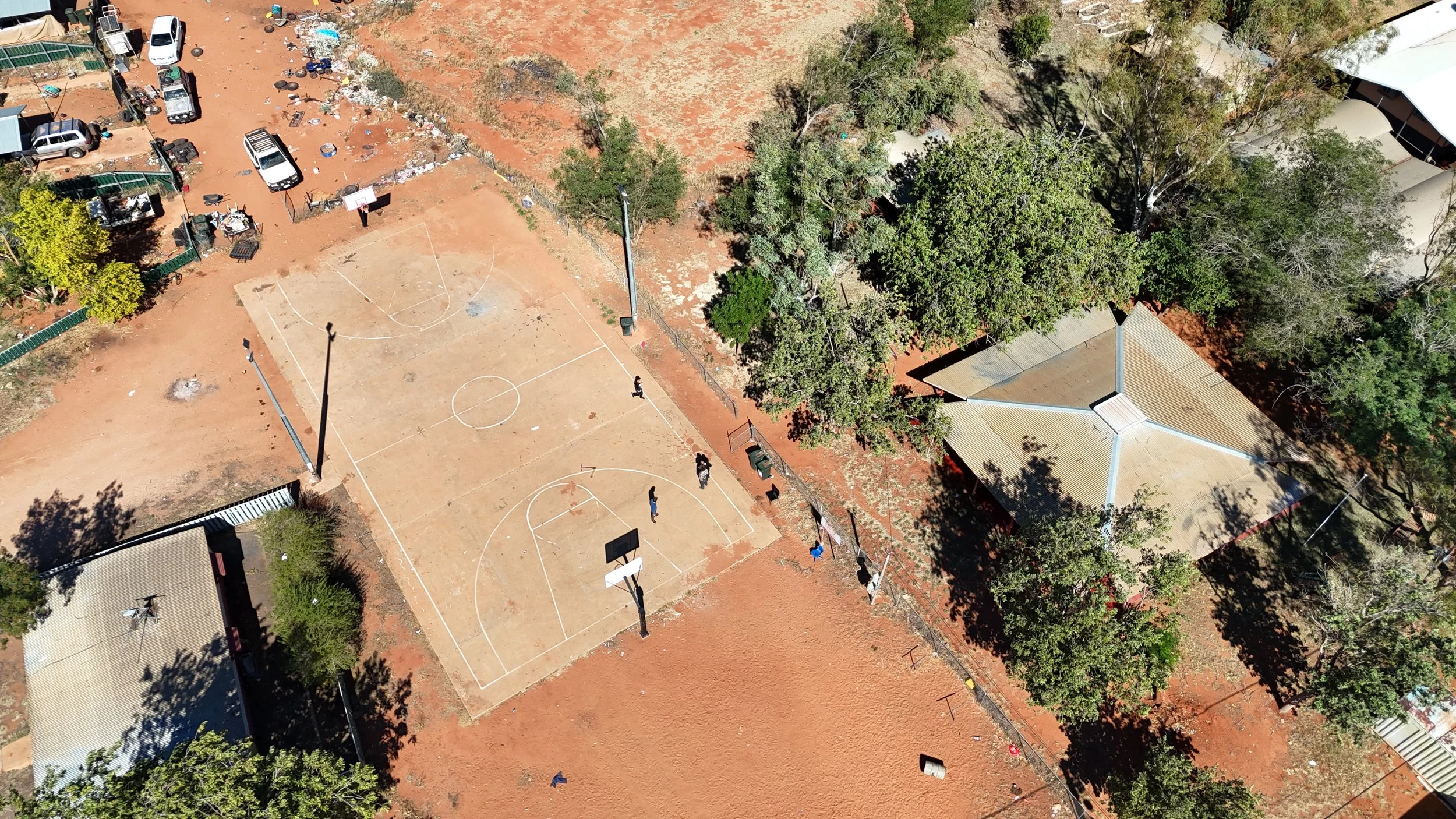 Aerial view of a basketball court surrounded by red earth, trees and other buildings