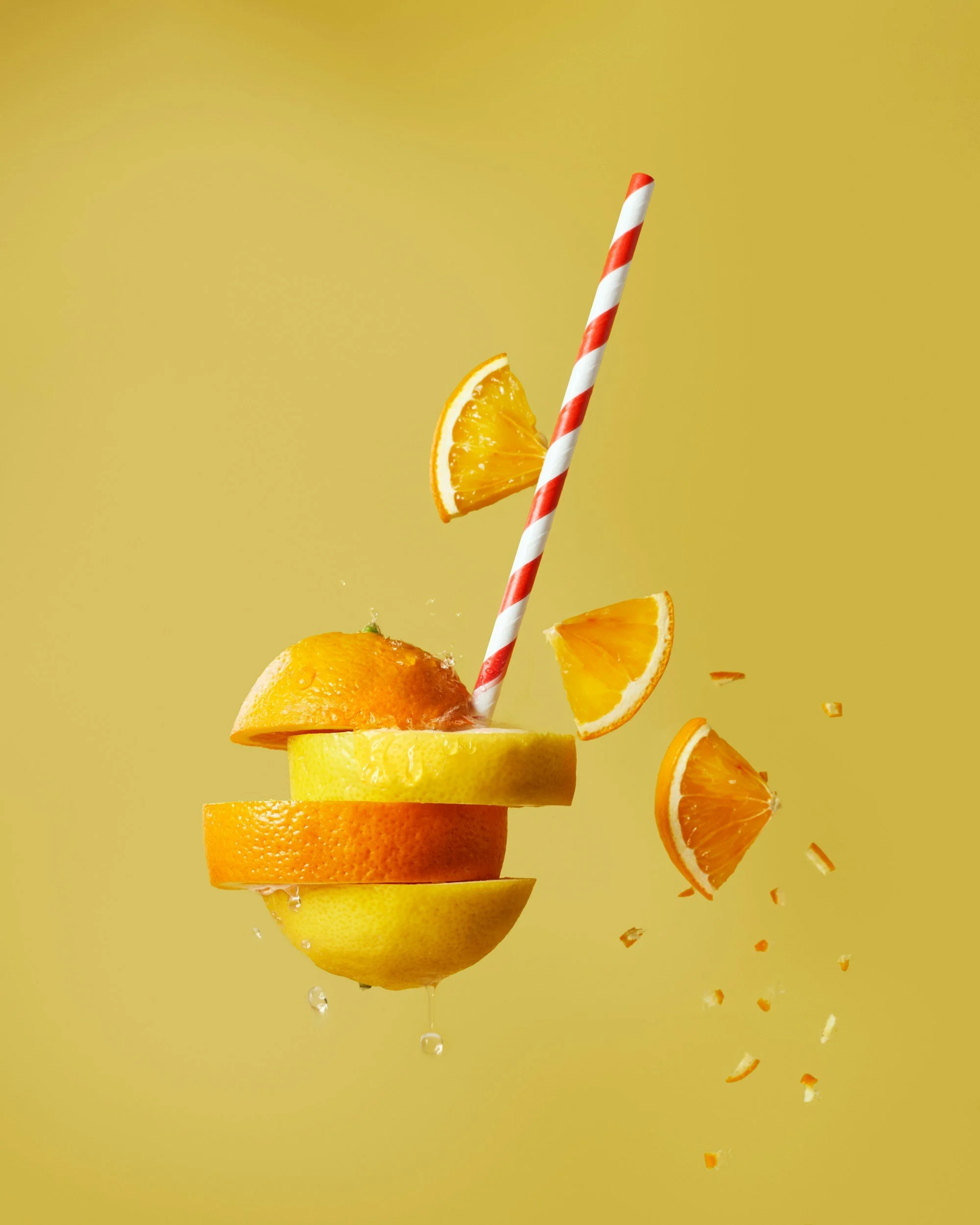 Orange and lemon slices arranged in a floating, stacked manner with a striped straw and orange wedges, against a yellow background.