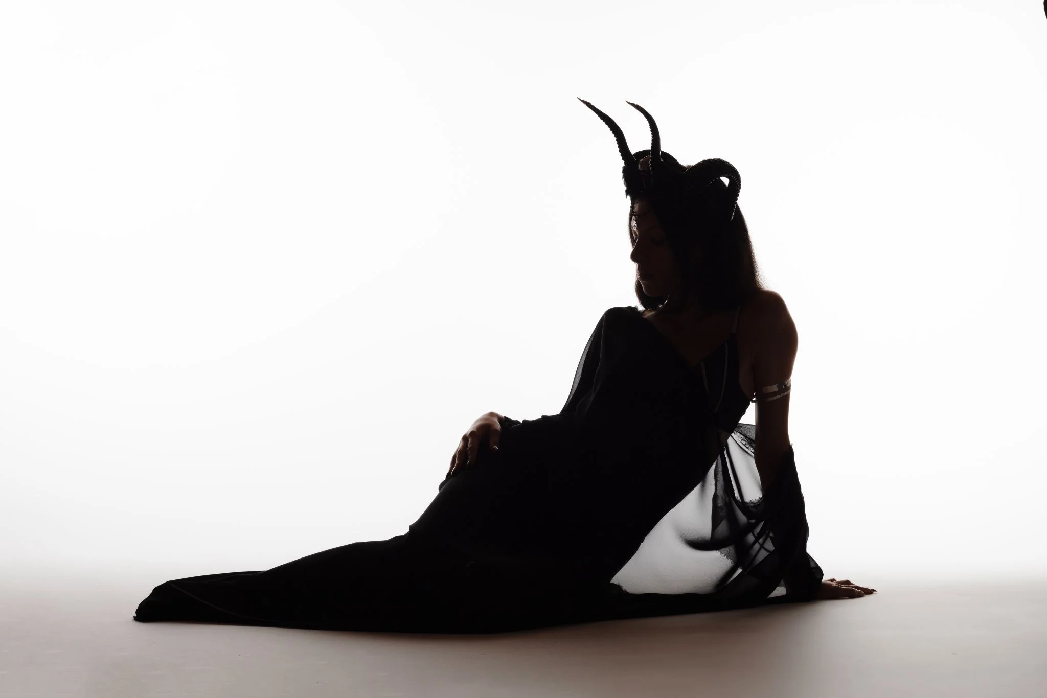 Silhouette einer Person mit Hörnern auf dem Kopf, sitzt auf dem Boden, vor weißem Hintergrund, dunkle Kleidung, weibliche Figur, im Schatten.