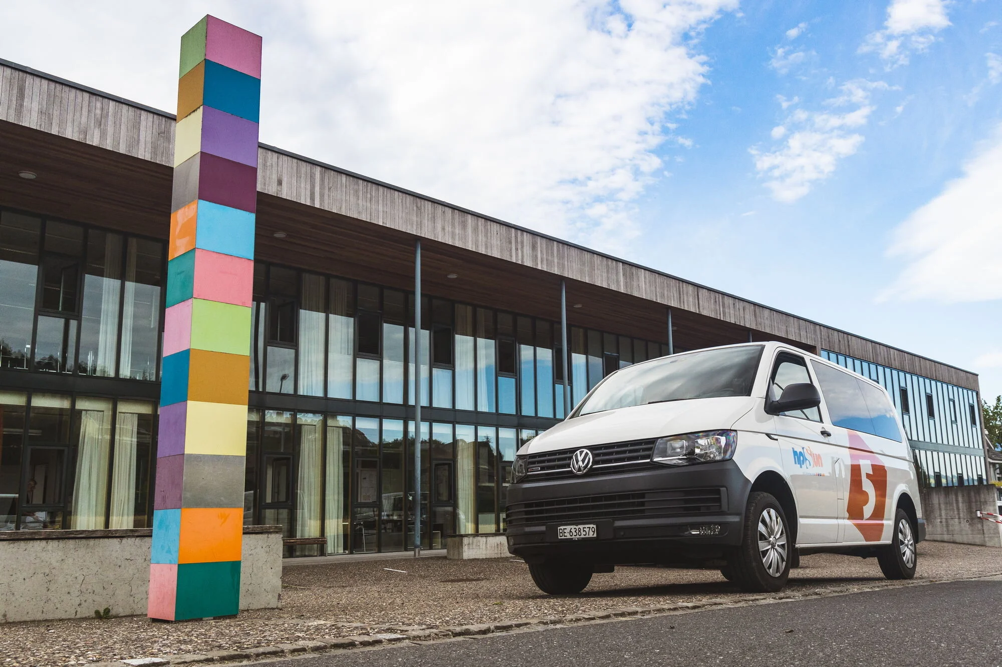 Ein weißer Volkswagen-Bus steht vor einem modernen Gebäude mit großen Glasfenstern und einer mehrfarbigen Säule. Das Gebäude hat eine Holzverkleidung und eine überdachte Terrasse.