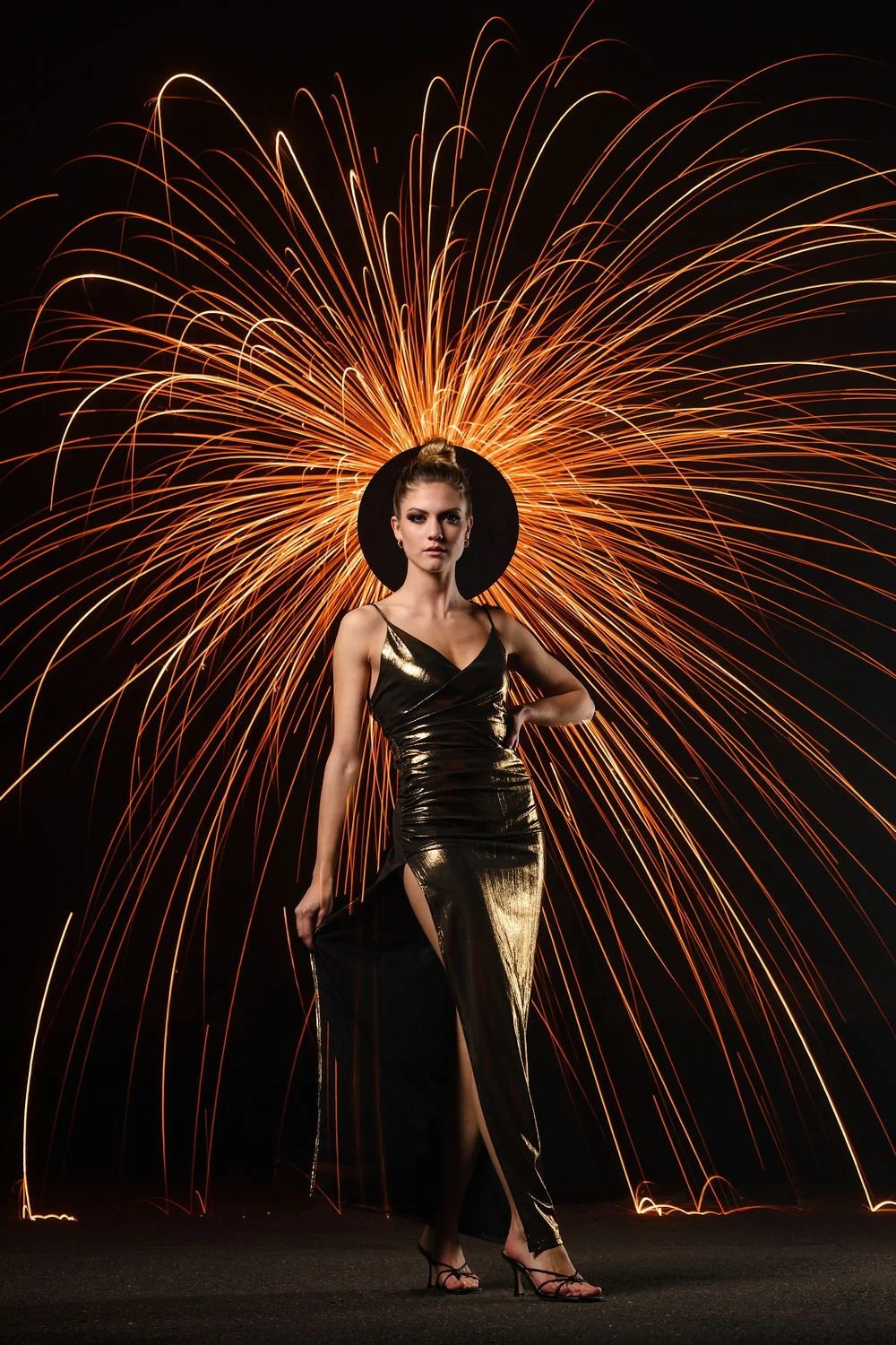 Frau in eleganter schwarzer Gold glänzender Dress mit hoher Schlitz, schwarzer Hut, vor einer Explosion aus Funken und Licht. Urbanes, festliches Ambiente.