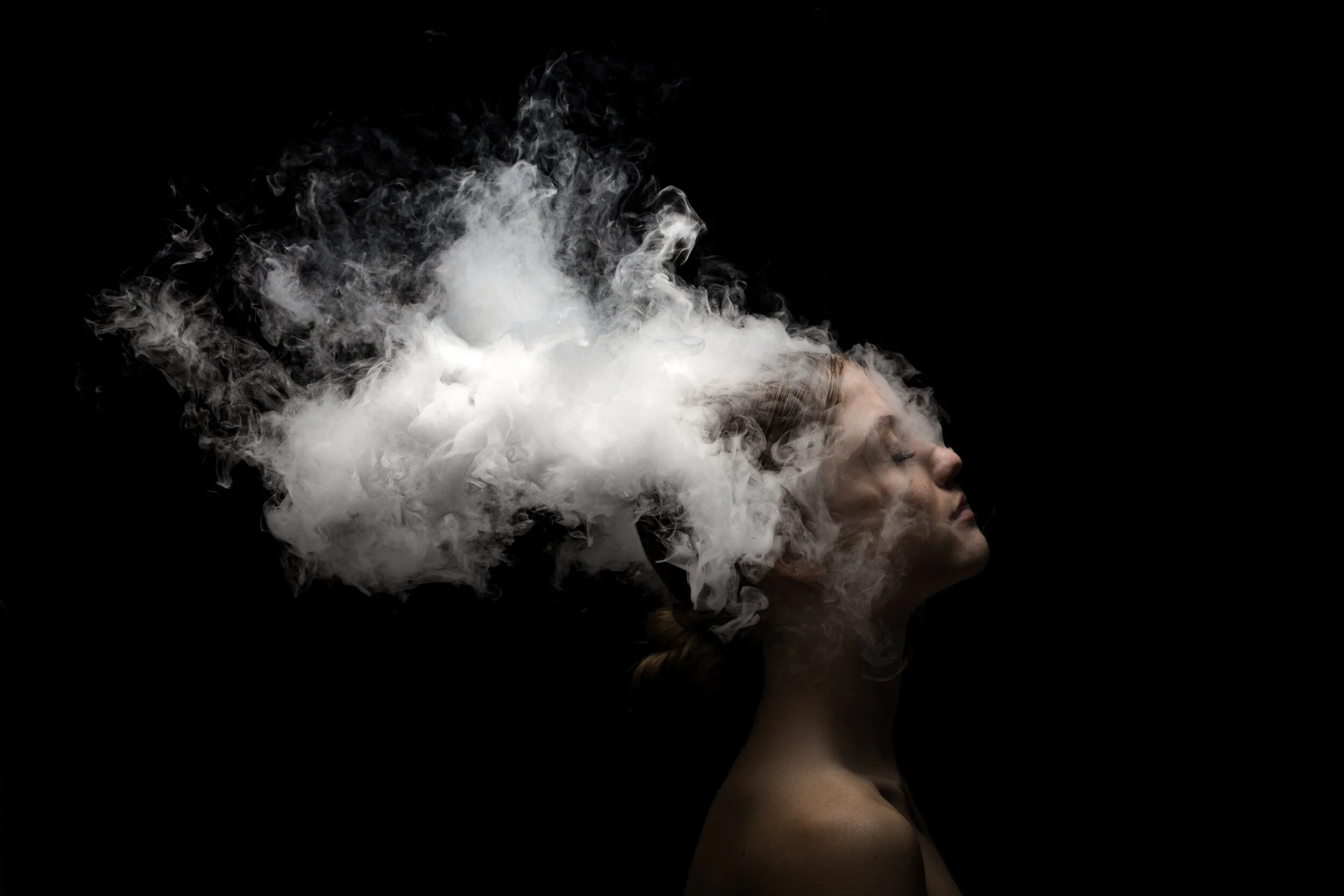 Porträt einer Frau mit geschlossenen Augen, deren Kopf von weißen Rauchschwaden umgeben ist, auf schwarzem Hintergrund.