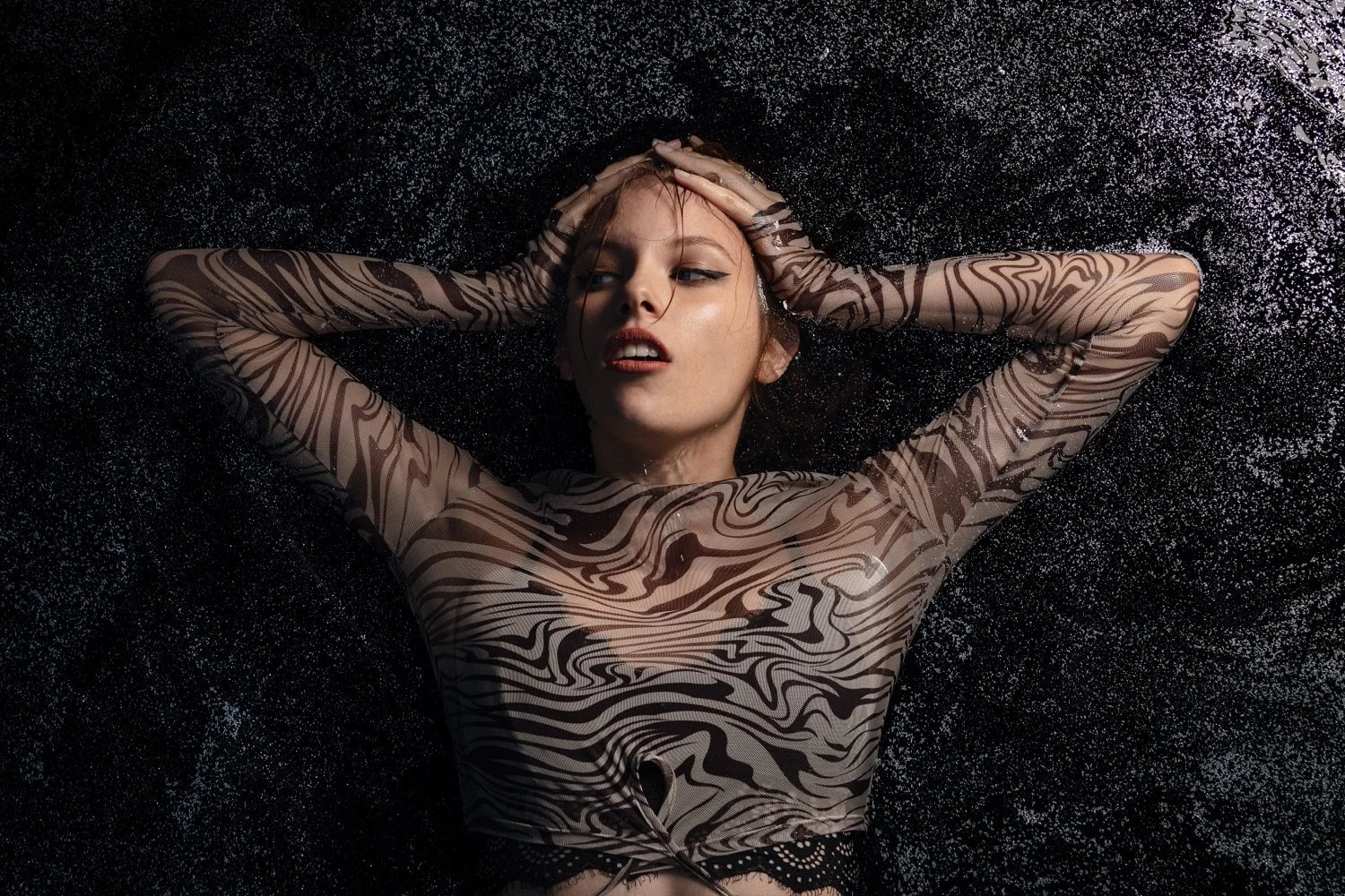 Junge Frau liegt auf schwarzem, glitzerndem Hintergrund, trägt durchsichtigen, langärmeligen Stoff mit Zebramuster, sich die Haare mähnend.
