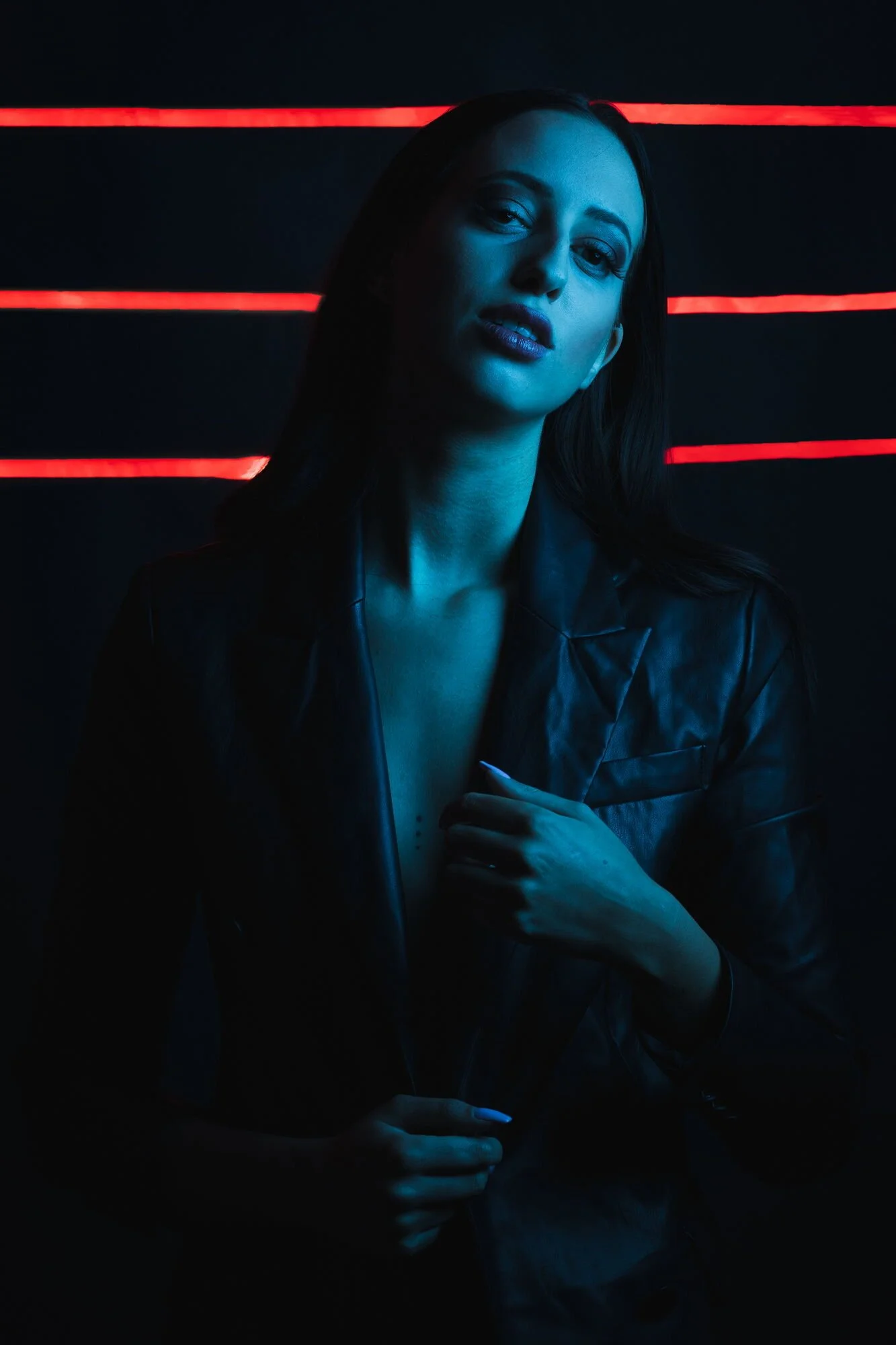 Eine dunkel beleuchtete Frau trägt eine schwarze Lederjacke, hinter ihr sind rote Neonlinien sichtbar, ihre Haut ist bluish-gefärbt, und sie schaut in die Kamera.