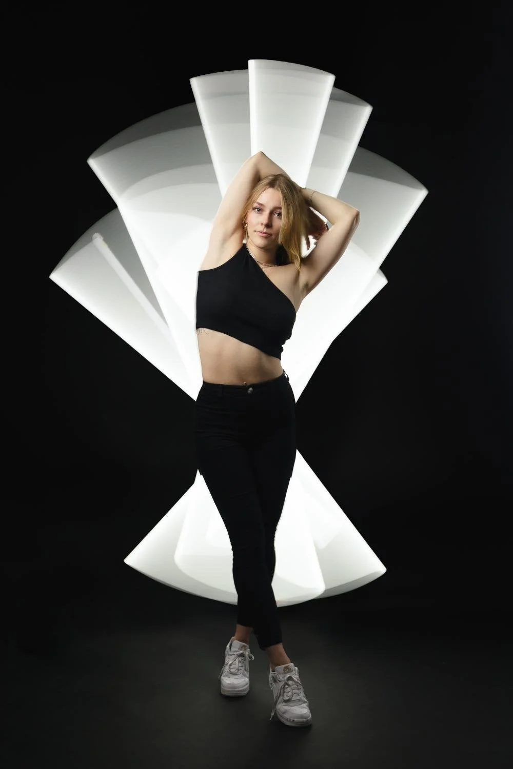 Junge Frau trägt schwarzes Top und schwarze Hosen, steht vor einer schwarzen Hintergrund mit weißen Lichtspuren im Hintergrund.