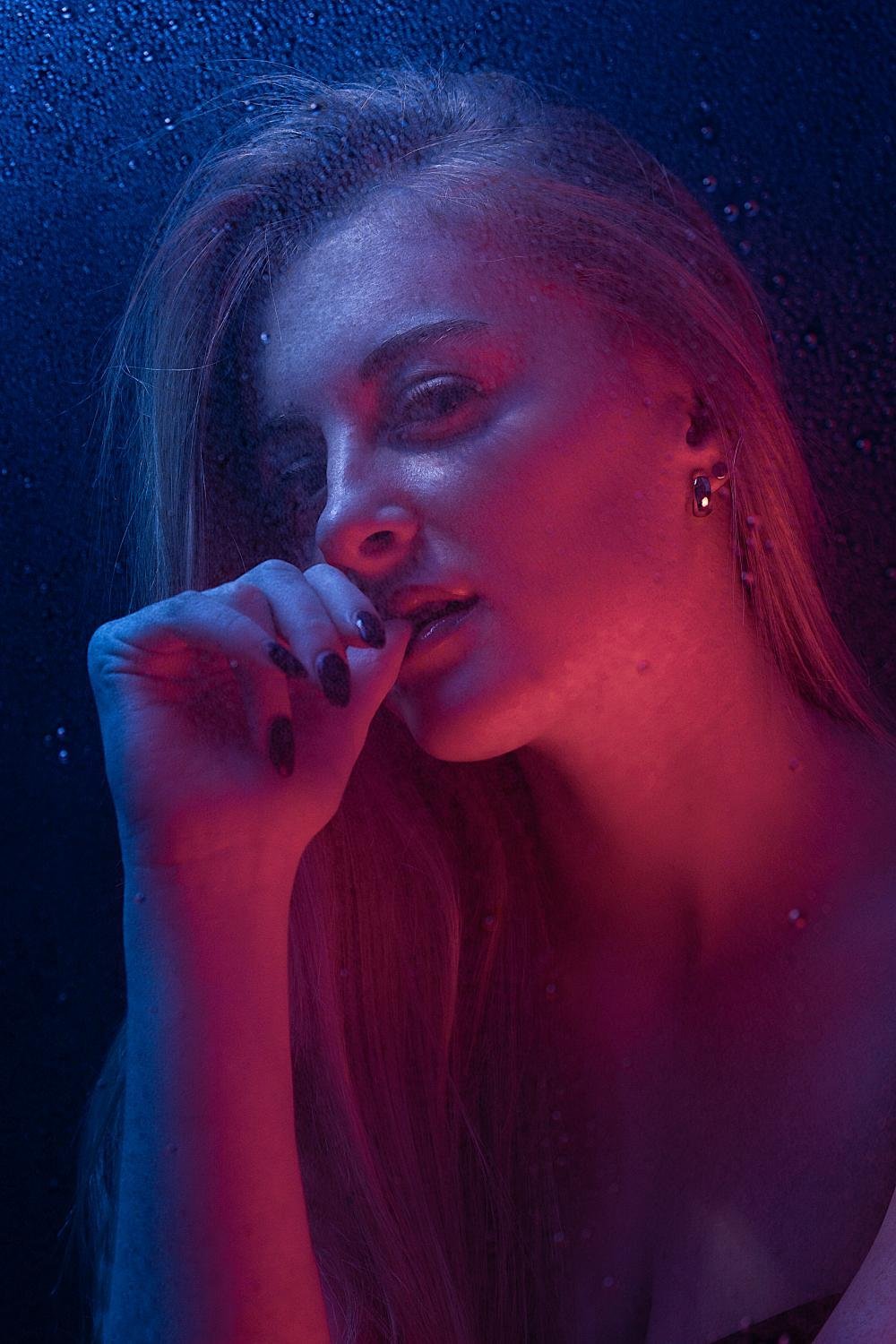 Porträt einer jungen Frau mit langem Haar, die in dunkler Umgebung mit buntem Neonlicht fotografiert wurde, die Hand vor den Mund hält, dunkle Nägel und Schmuck trägt.