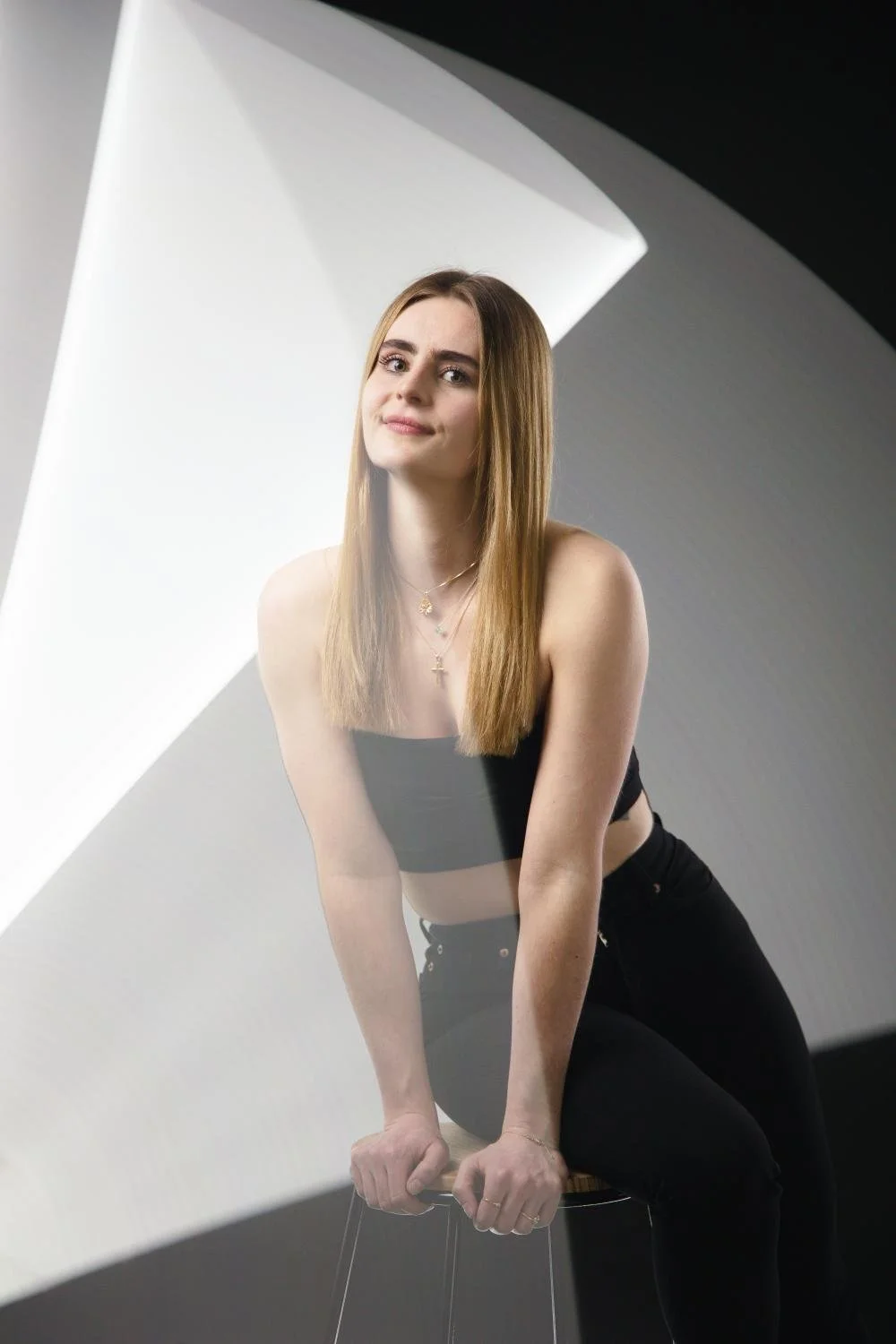 Ein junges Mädchen mit longem, hellbraunem Haar sitzt auf einem Barhocker in einem stilvollen, modern gestalteten Raum. Sie trägt ein schwarzes Crop-Top, schwarze Hose und mehrere Halsketten. Im Hintergrund ist eine große Lichtquelle, die den Raum gu