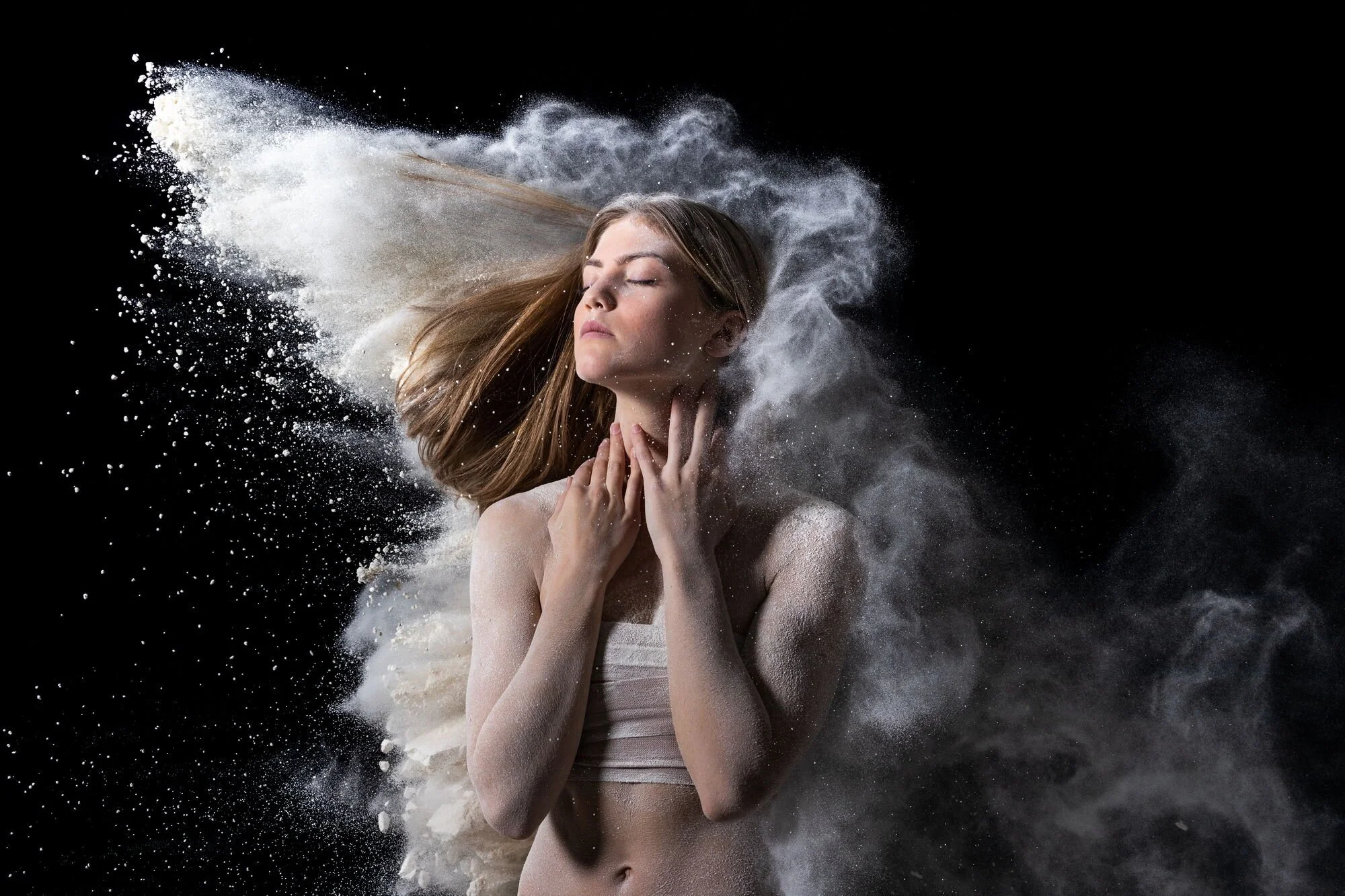 Junge Frau mit geschlossenen Augen und entspanntem Gesicht, umgeben von Mehl oder Staub, der sich in der Luft verteilt, vor einem schwarzen Hintergrund.
