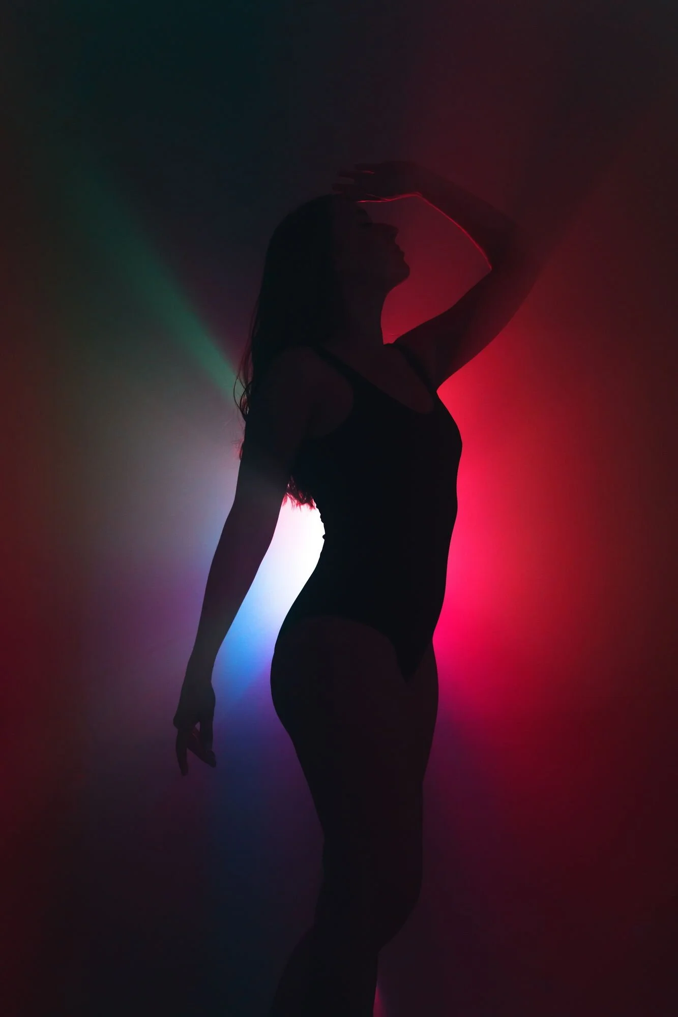 Silhouette einer Frau in dunkler Kleidung vor buntem Hintergrund, die mit der Hand den Kopf berührt, in einer künstlerischen, lichtspielartigen Pose.