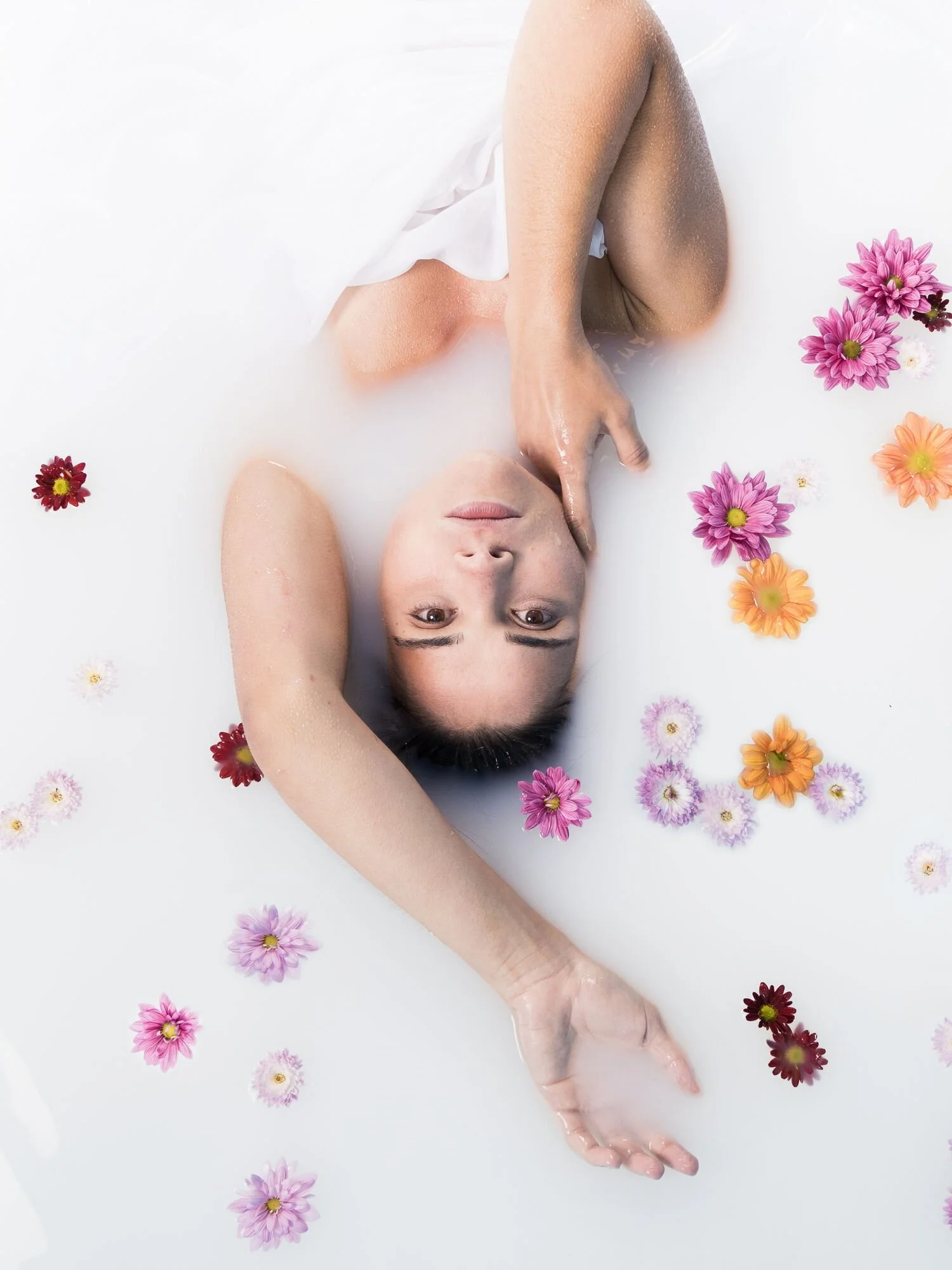 Frau liegt in Milchbad mit Blumen, entspannt, Blick nach oben, umgeben von bunten Blumen.