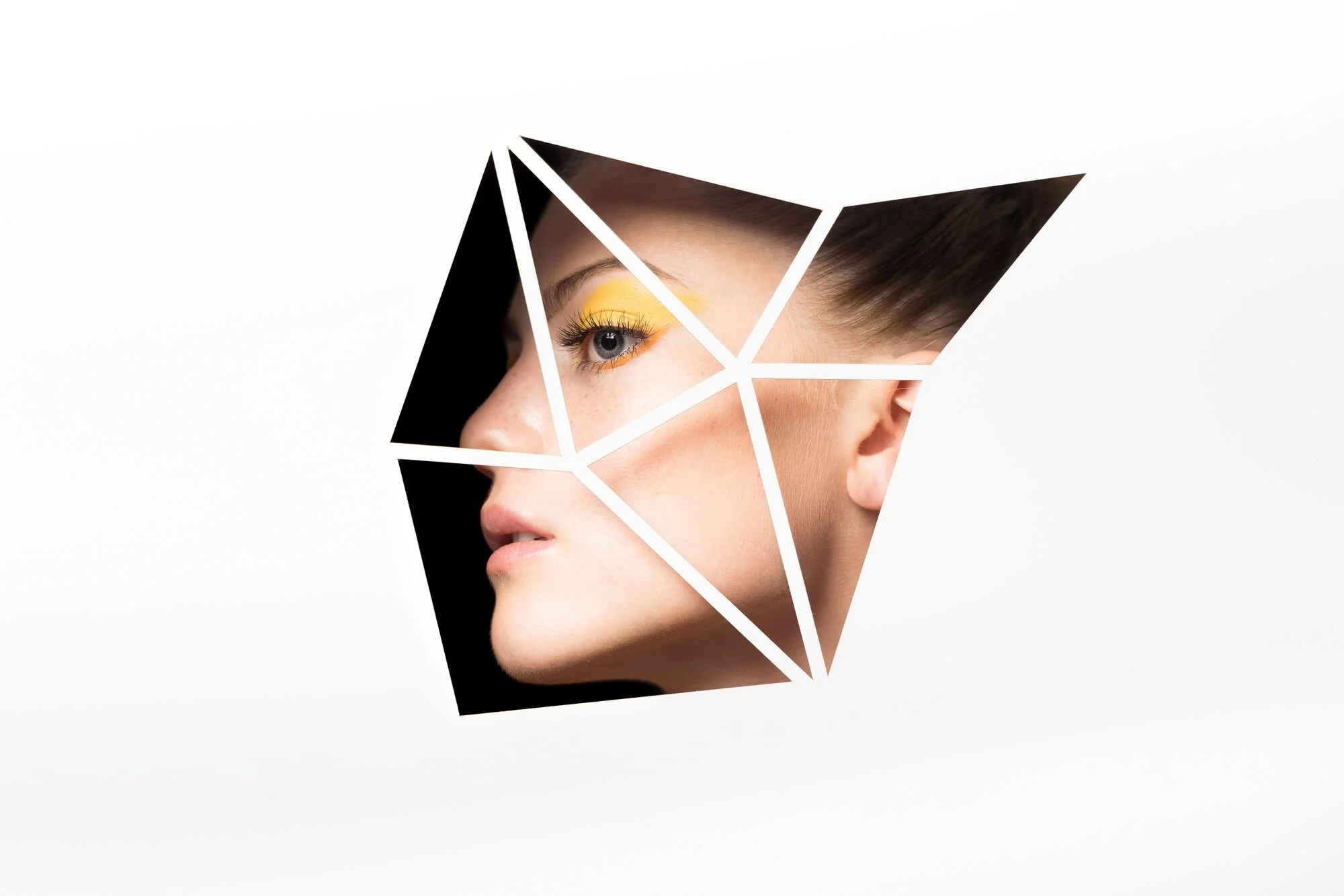 Kunstvoll zugeschnittenes Porträt einer jungen Frau mit gelbem Augen-Make-up, sichtbar durch geometrische Schnittmuster auf einer weißen Fläche.