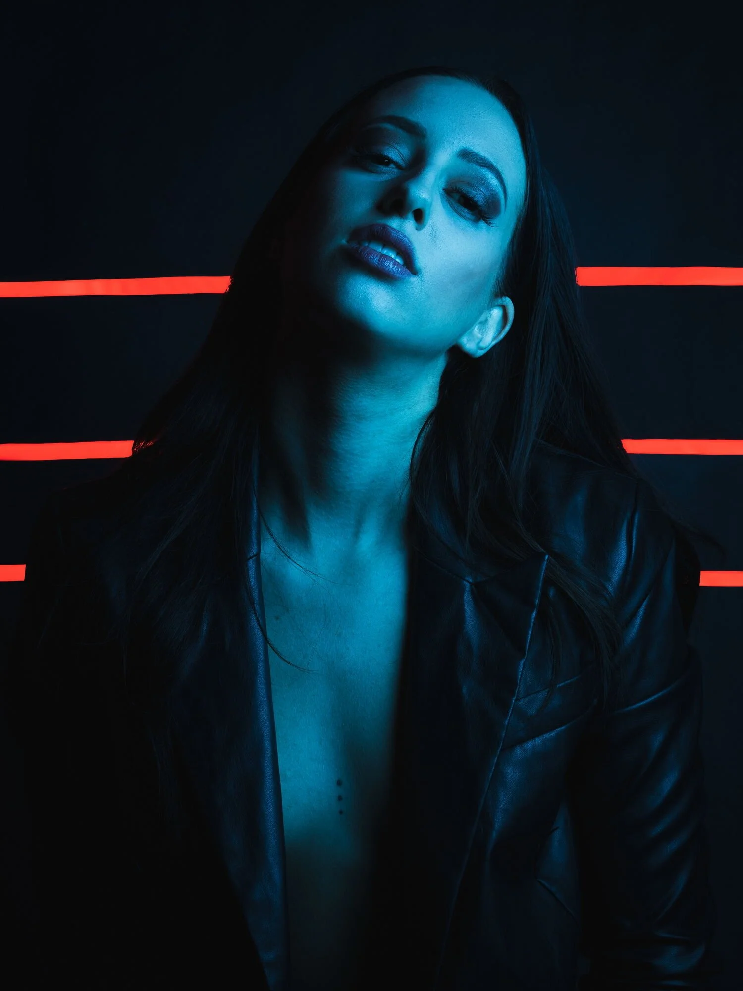 Junge Frau mit dunklen Haaren, trägt einen schwarzen Lederjacke vor einem schwarzen Hintergrund mit roten Linien, beleuchtet in blauer Lichtstimmung.