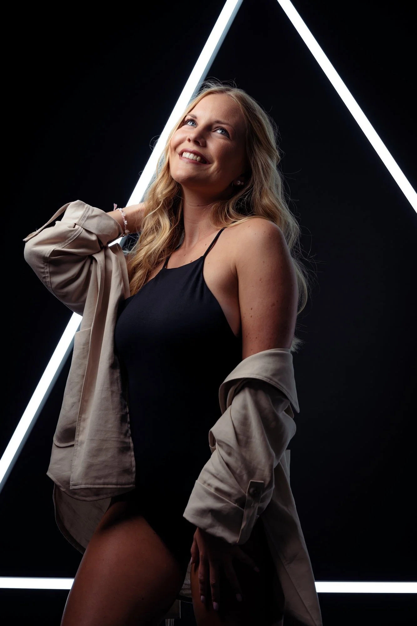 Lächelnde junge Frau mit langem, gewelltem Haar trägt schwarzen Spaghettiträger-Body und beige Jacke, im Hintergrund neonbeleuchtete Dreiecke auf schwarzem Hintergrund.