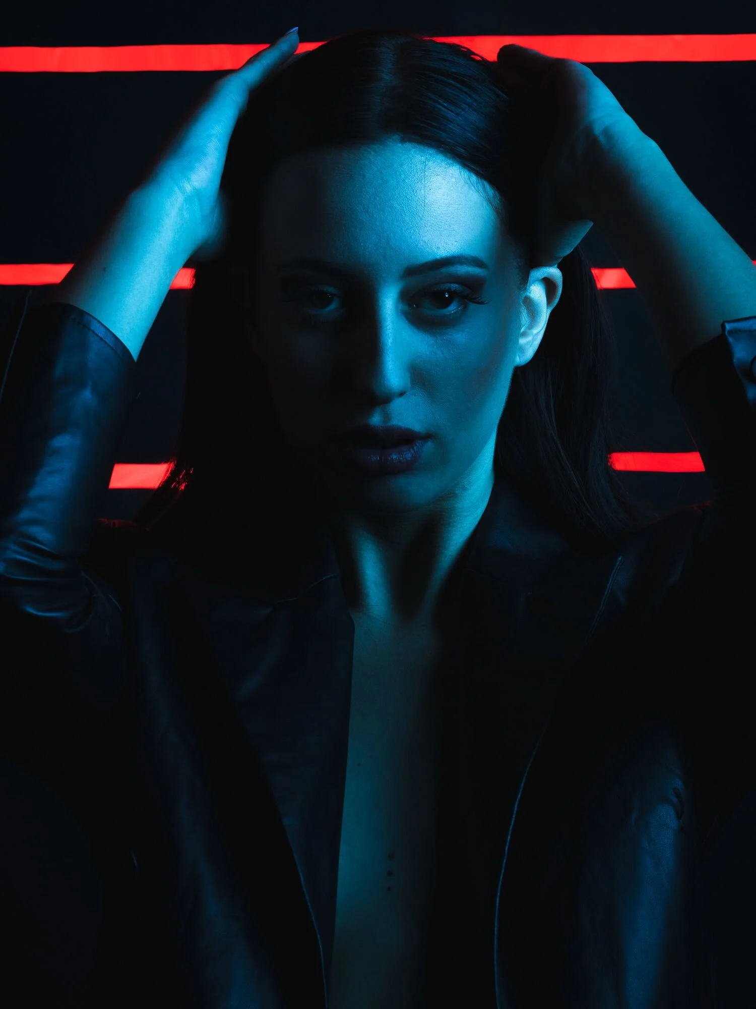 Eine junge Frau mit dunklen Haaren in einem schwarzen Leder-Outfit, die in einer dunklen Umgebung mit roten Lichtstreifen posiert.