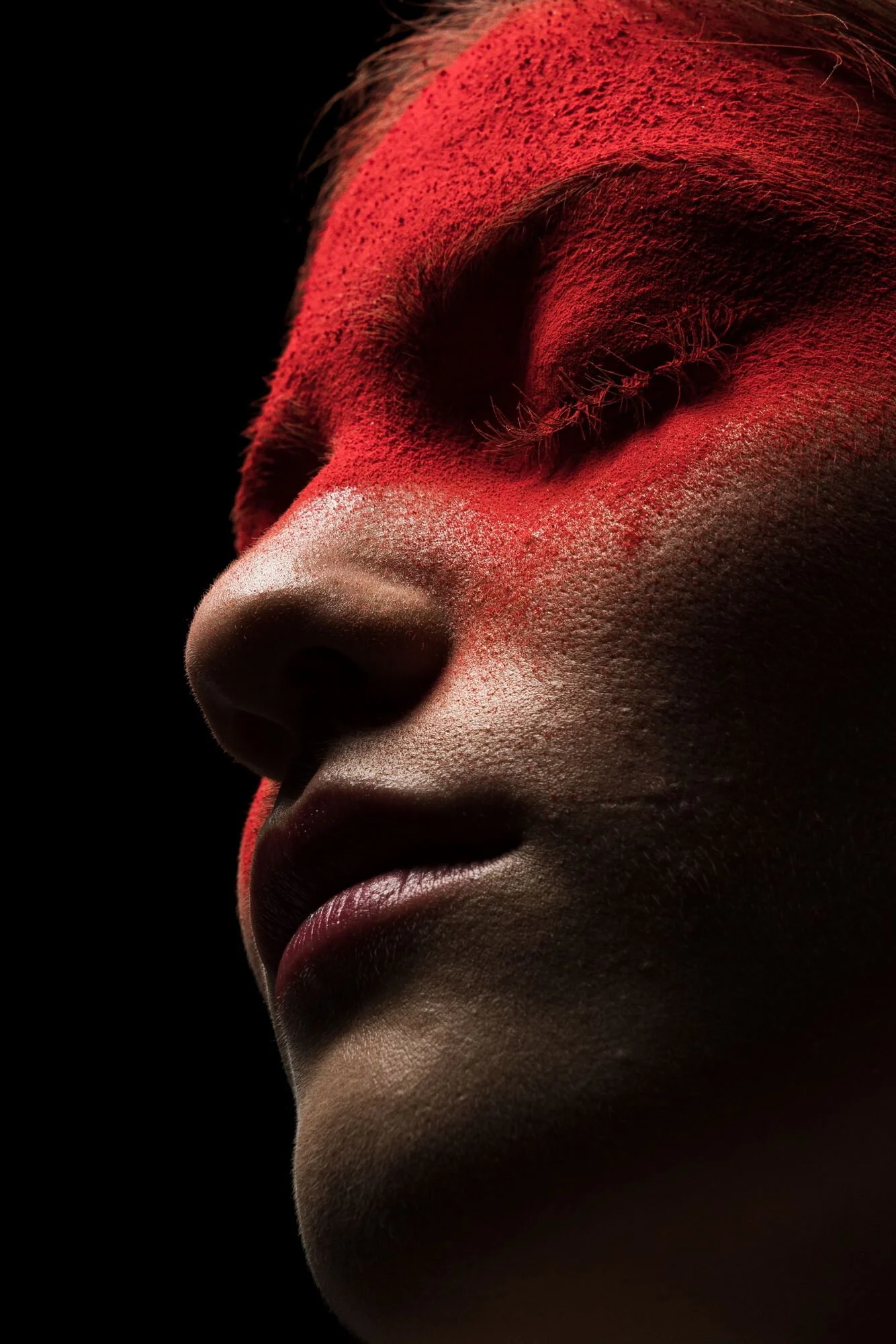 Nahaufnahme eines Gesichts mit roter Farbe auf der Stirn und Wange, geschlossene Augen, dunkler Hintergrund.