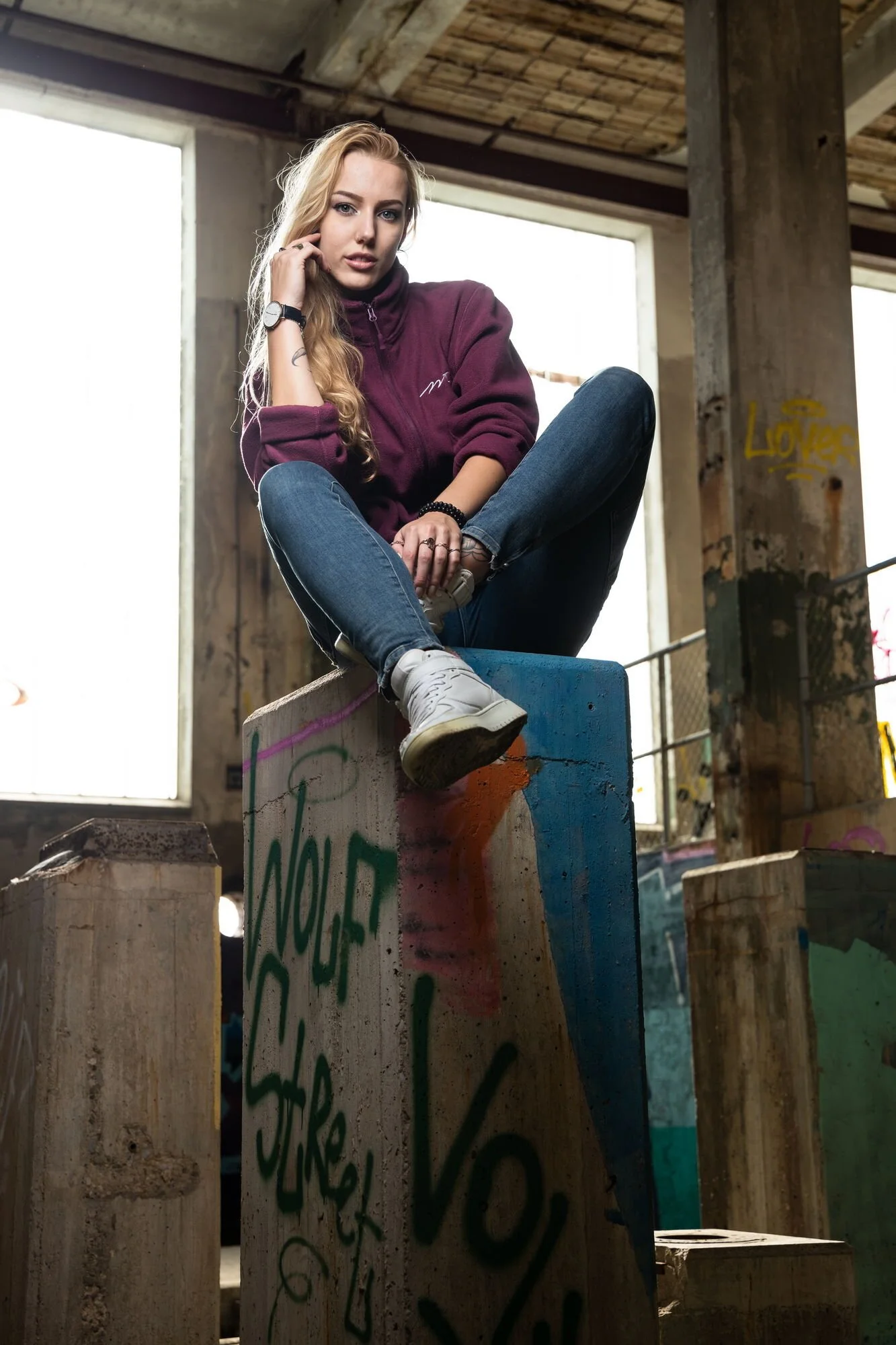 Junge Frau sitzt auf einem graffiti-bemalten Holzblock in einem alten, verfallenen Gebäude mit großen Fenstern und Graffiti an den Wänden.