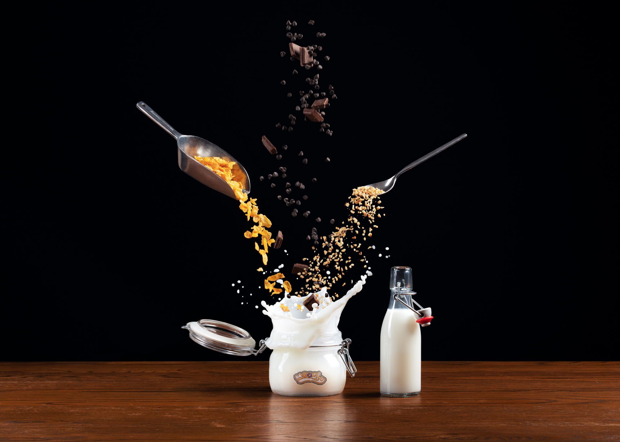 Milch, Cornflakes und Schokoladenstückchen fliegen in der Luft, während sie in einen offenen Behälter auf einem Holztisch fallen, mit einem schwarzen Hintergrund.