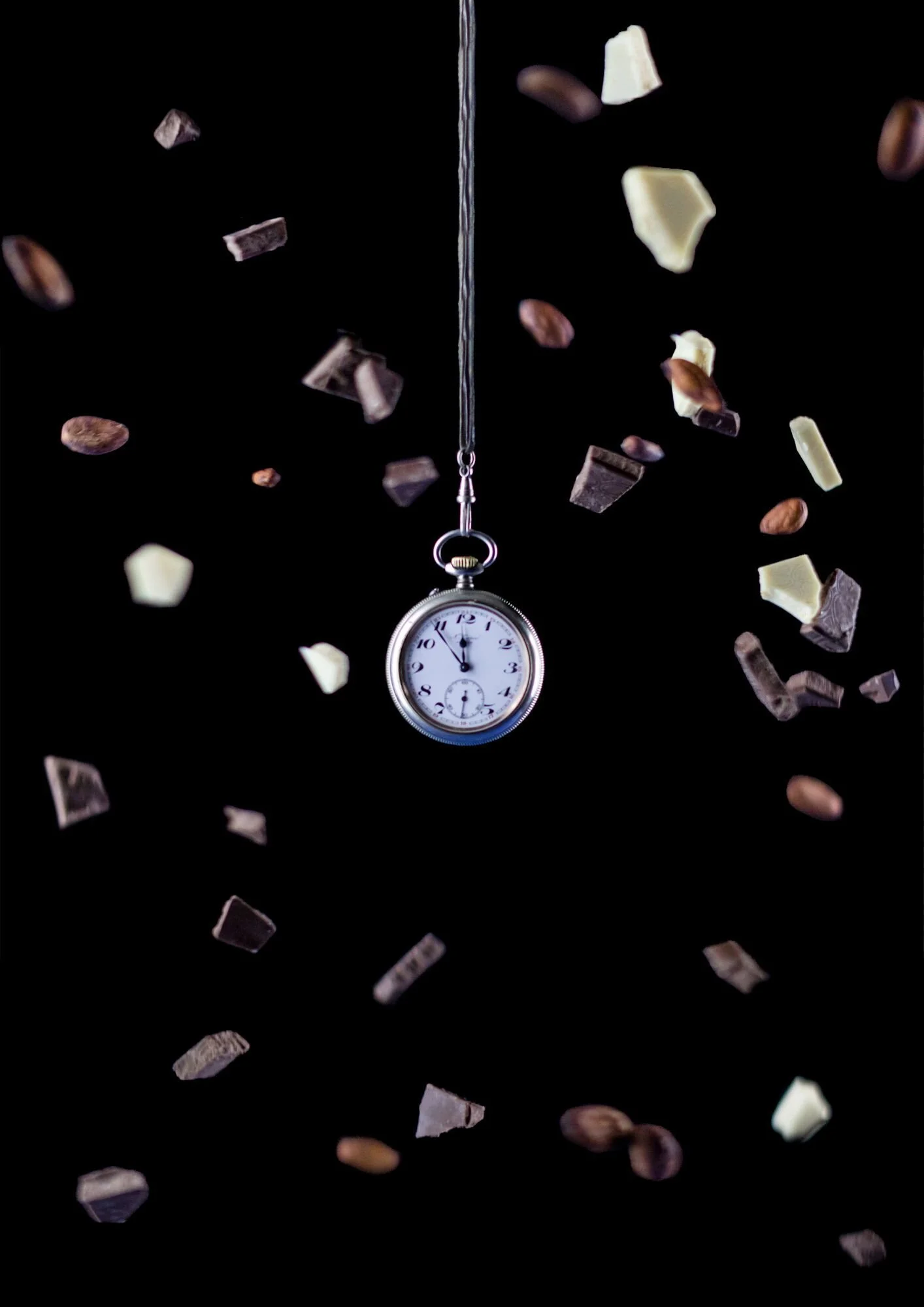 Eine Taschenuhr hängt an einem Schnur und umkreist von fallenden Schokoladenstücken und Nüssen vor einem schwarzen Hintergrund.