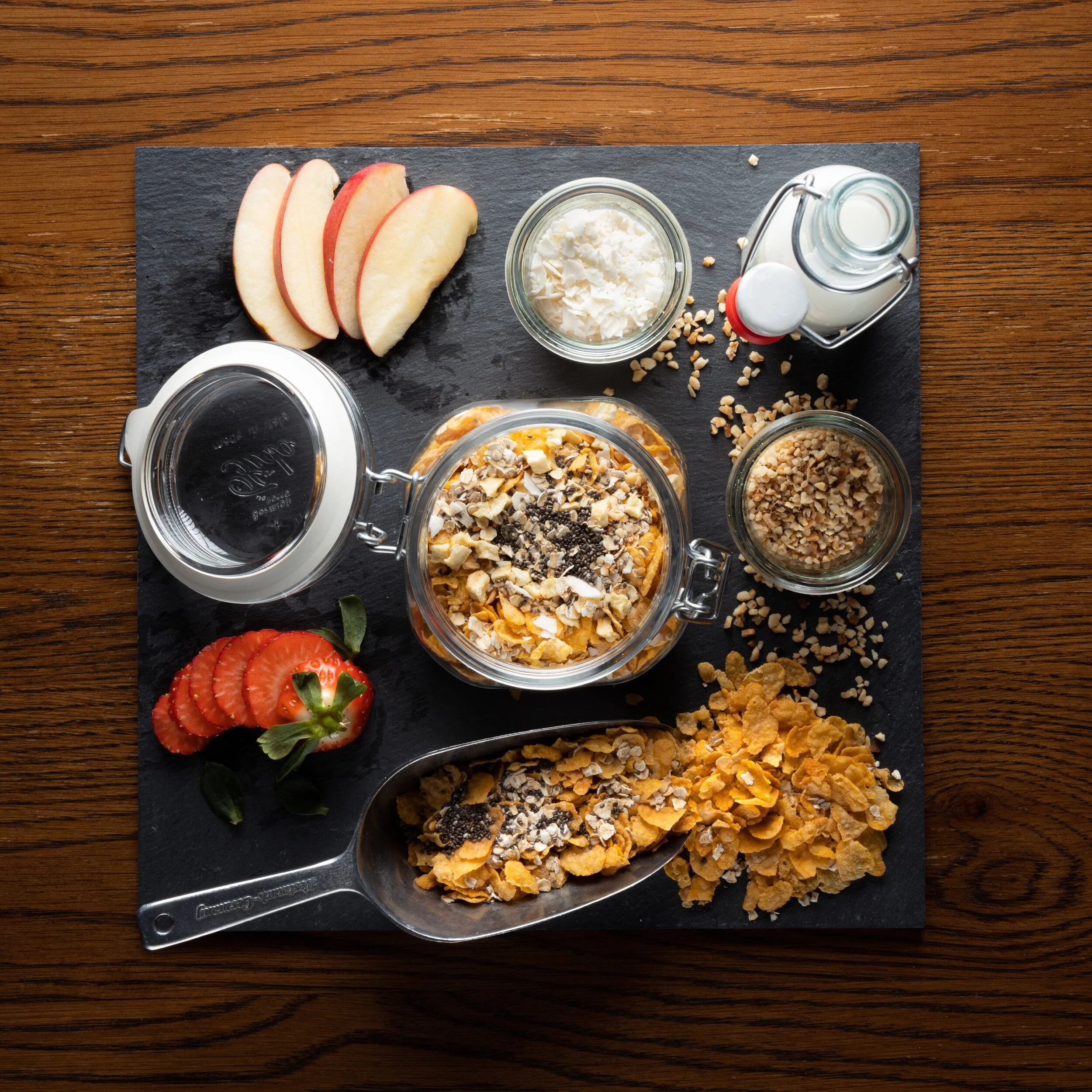 Verschiedene Zutaten für Müsli, darunter Apfelscheiben, Erdbeeren, Nüsse, Cornflakes, Joghurt, Milch und Haferflocken, auf einer schwarzen Platte auf einem Holztisch.