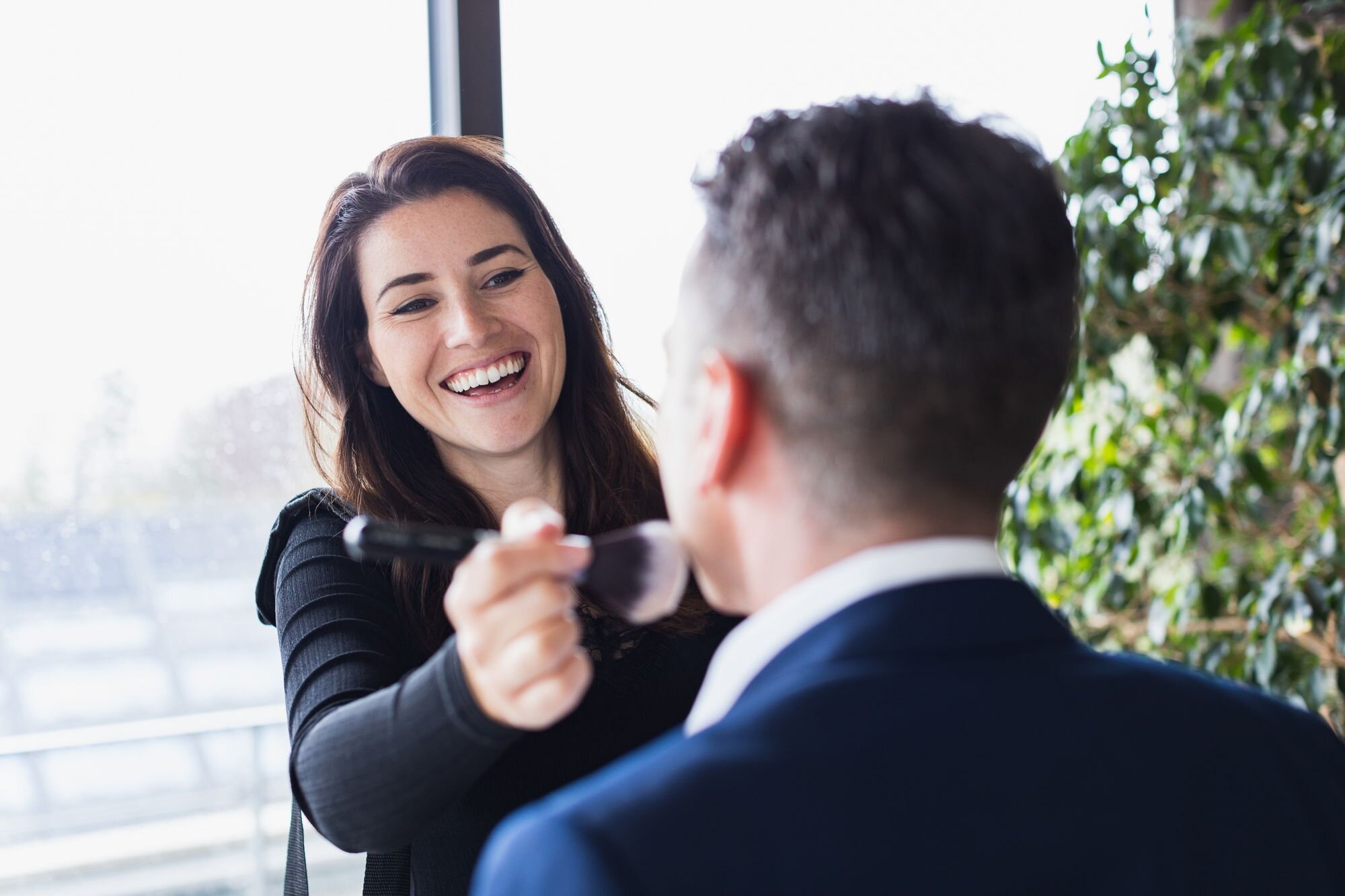 Eine lächelnde Frau beim Make-up bei einem Mann im Anzug.