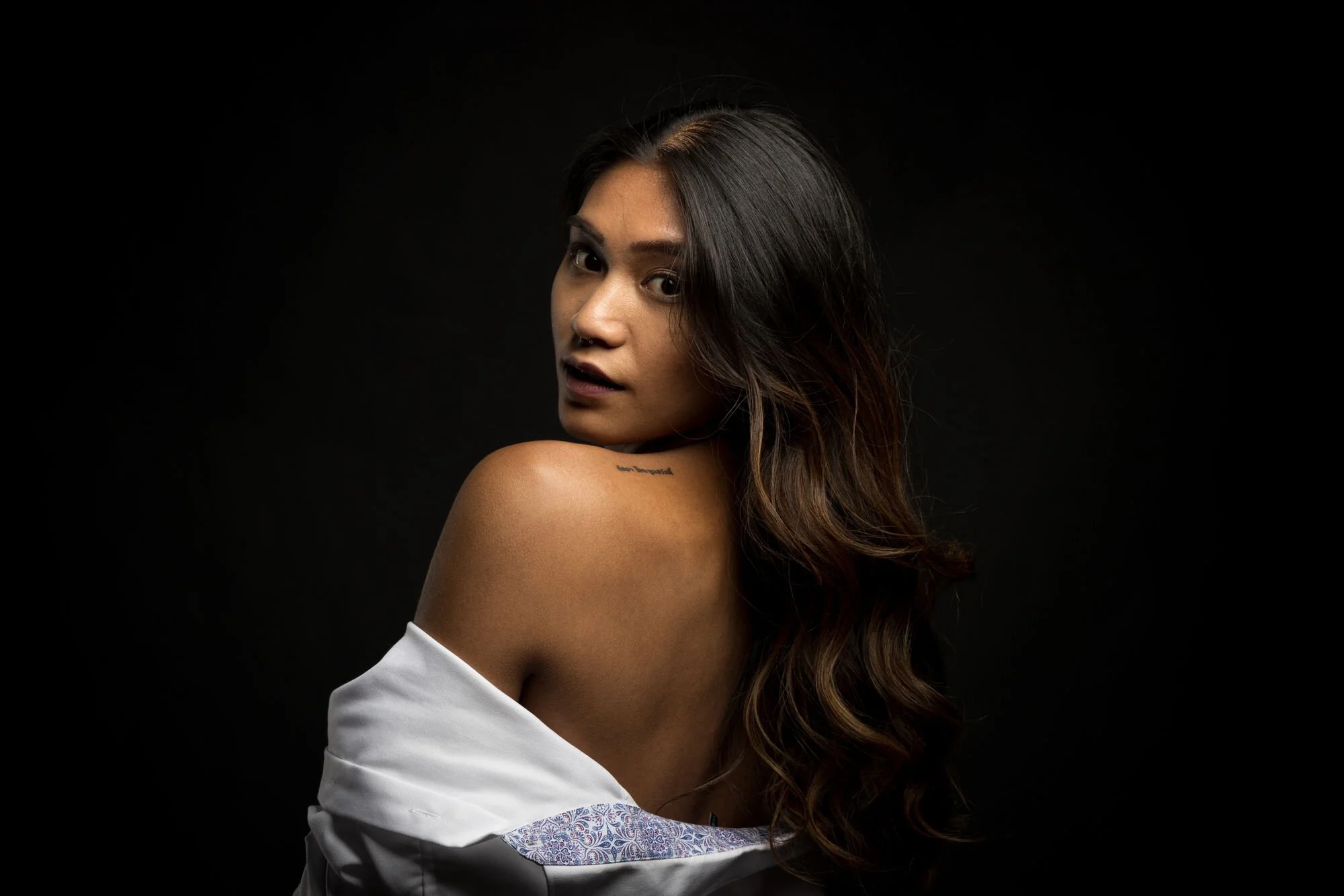 Junge Frau mit braunen, welligen Haaren, nacktem Schulter, in einem weißen Hemd gegen schwarzen Hintergrund.
