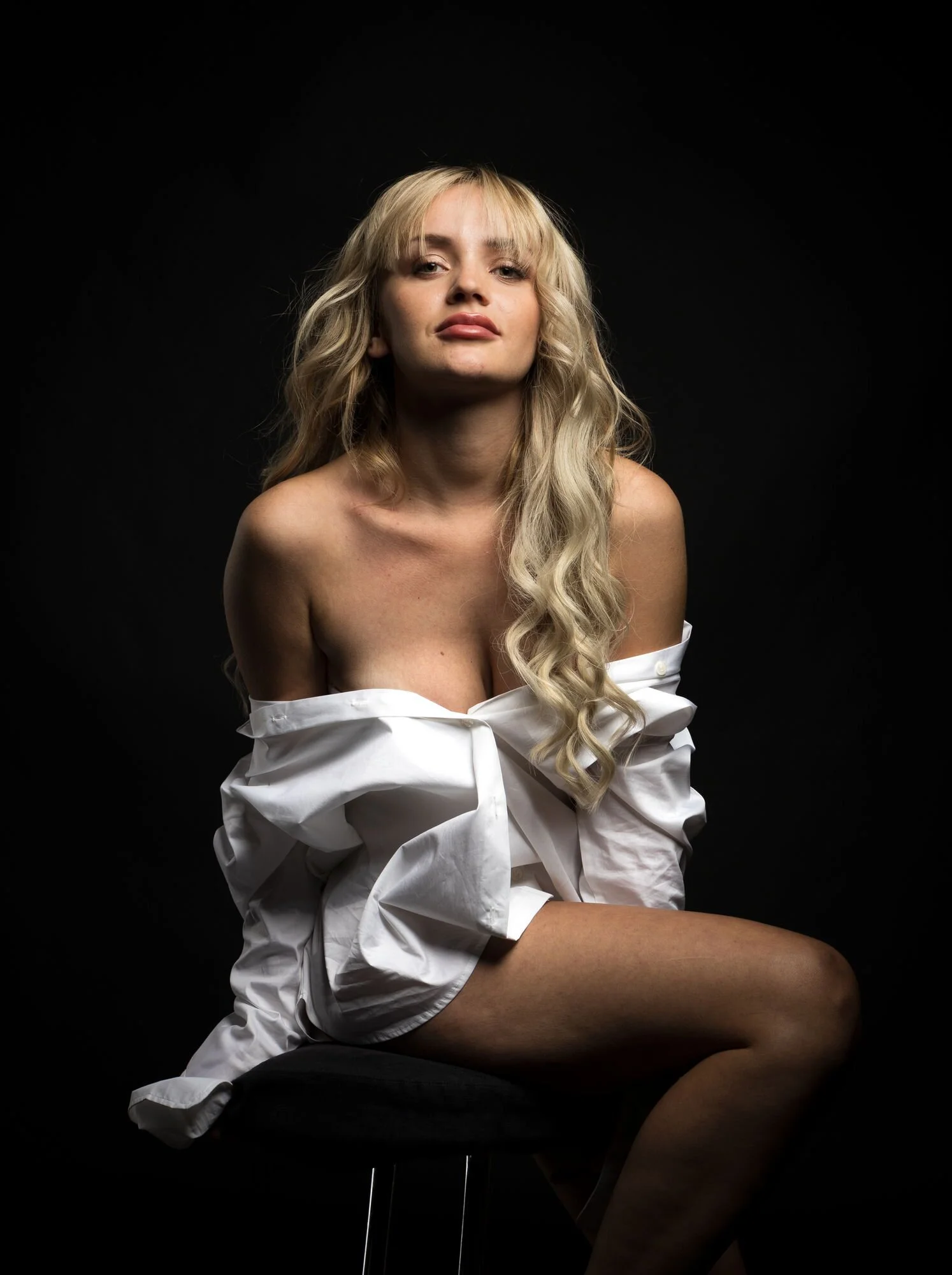 Eine junge Frau mit langen, lockigen blonden Haaren, bekleidet mit einem weißen Hemd, sitzt auf einem Stuhl vor schwarzem Hintergrund.