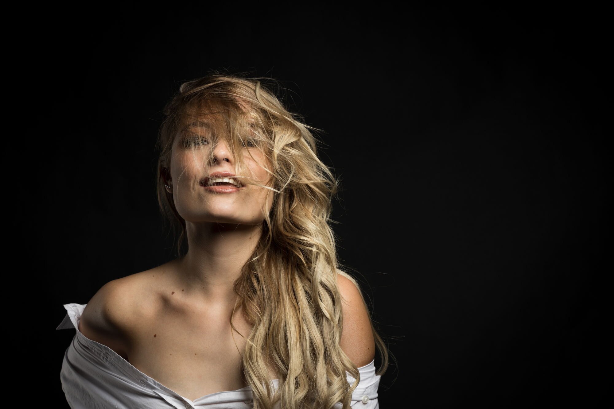 Junge Frau mit langen, lockigen Haaren und off-shoulder Shirt vor dunklem Hintergrund, lächelt leicht.