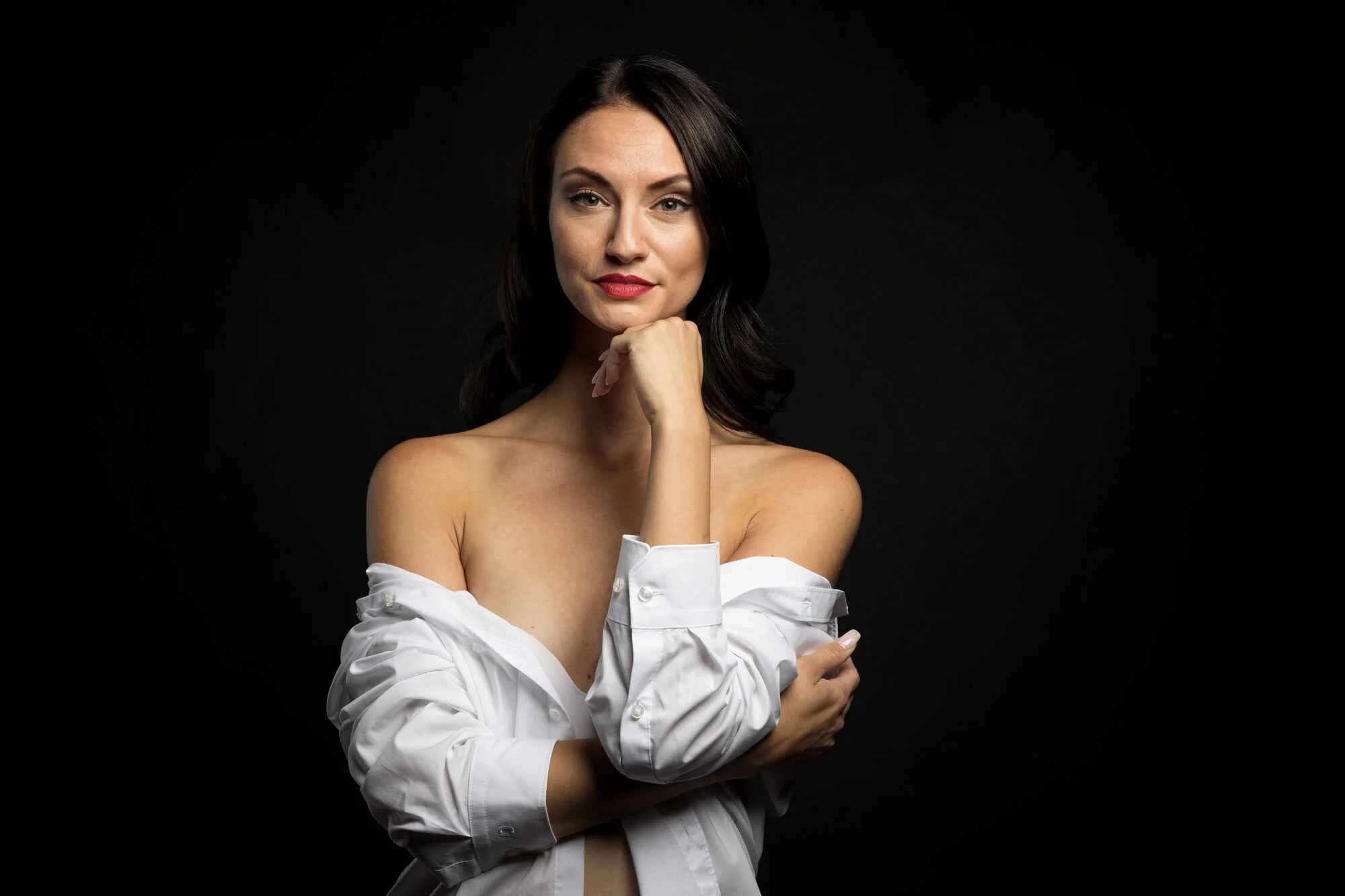 Junge Frau mit dunklen Haaren, nackt bis auf eine offene weiße Hemd, die lächelnd und mit einer Hand ihr Kinn stützt, vor schwarzem Hintergrund.