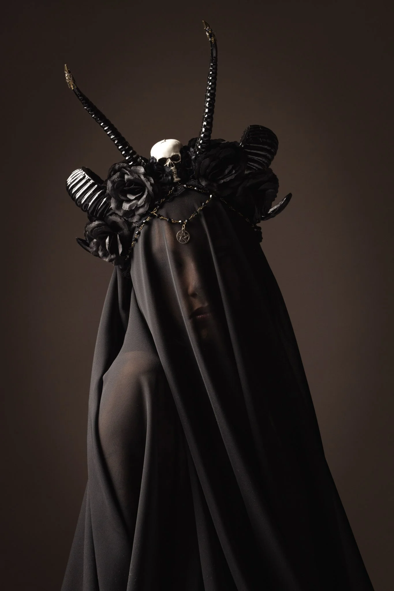 Junge Frau in dunklem, transparentem Schleier mit satanischem Symbol, schwarzer Rosenkranz, schwarzen Hörnern und dunkler Haut. Verschleierter Gesichtsausdruck, dunkler Hintergrund.