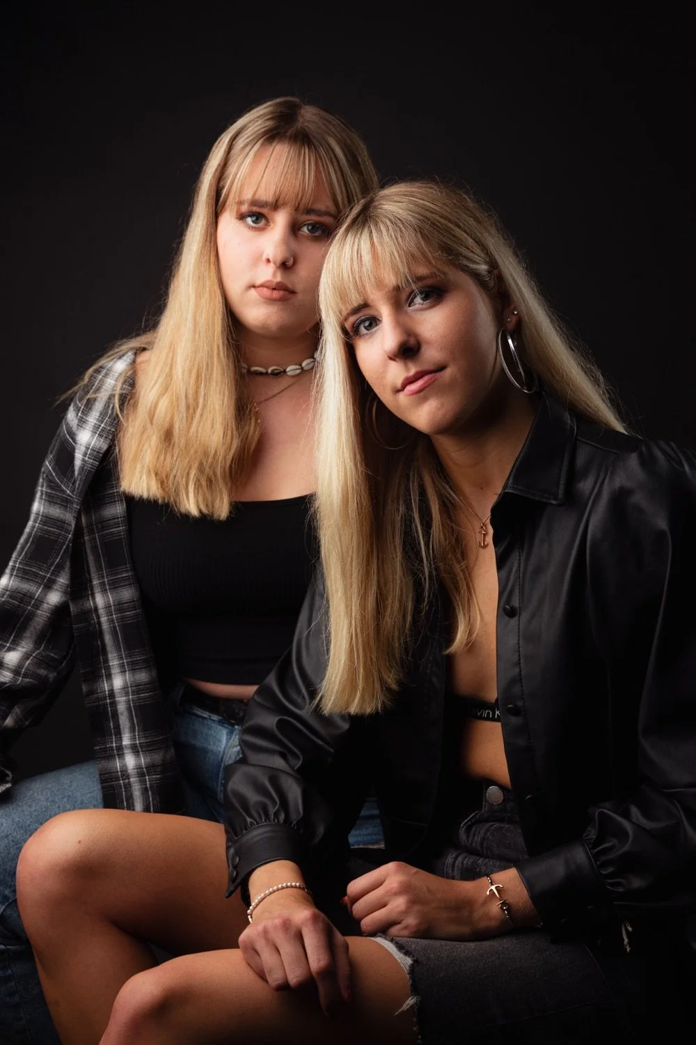 Zwei junge Frauen sitzen vor einem schwarzen Hintergrund, schauen ernst in die Kamera, tragen lässige Kleidung und haben blonde Haare.