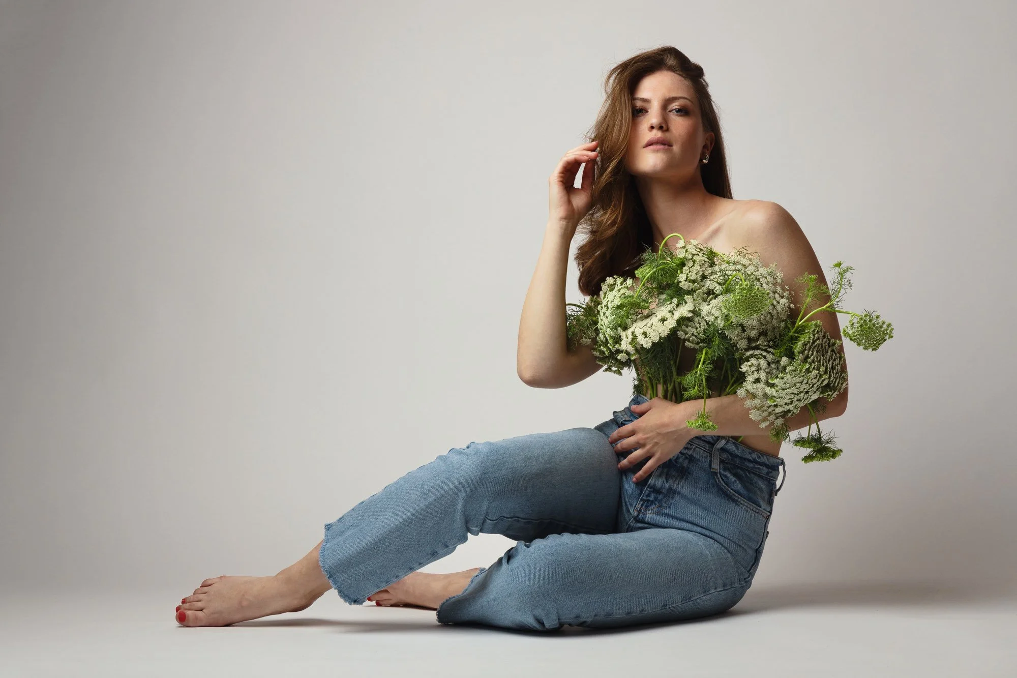 Junge Frau sitzt auf dem Boden, hält einen großen Blumenstrauß aus weißen und grünen Blumen, trägt Jeans und ist nackt obenrum, mit lockigem braunem Haar, vor hellem Hintergrund.