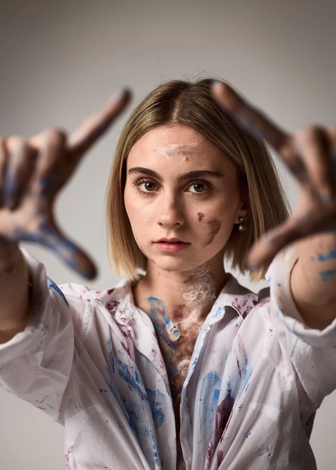Junge Frau mit Malfarbe auf Gesicht, Hals und Kleidung, steigt auf, Blick direkt in Kamera, trägt weiße Hemd, kurze blonde Haare, neutraler Hintergrund.