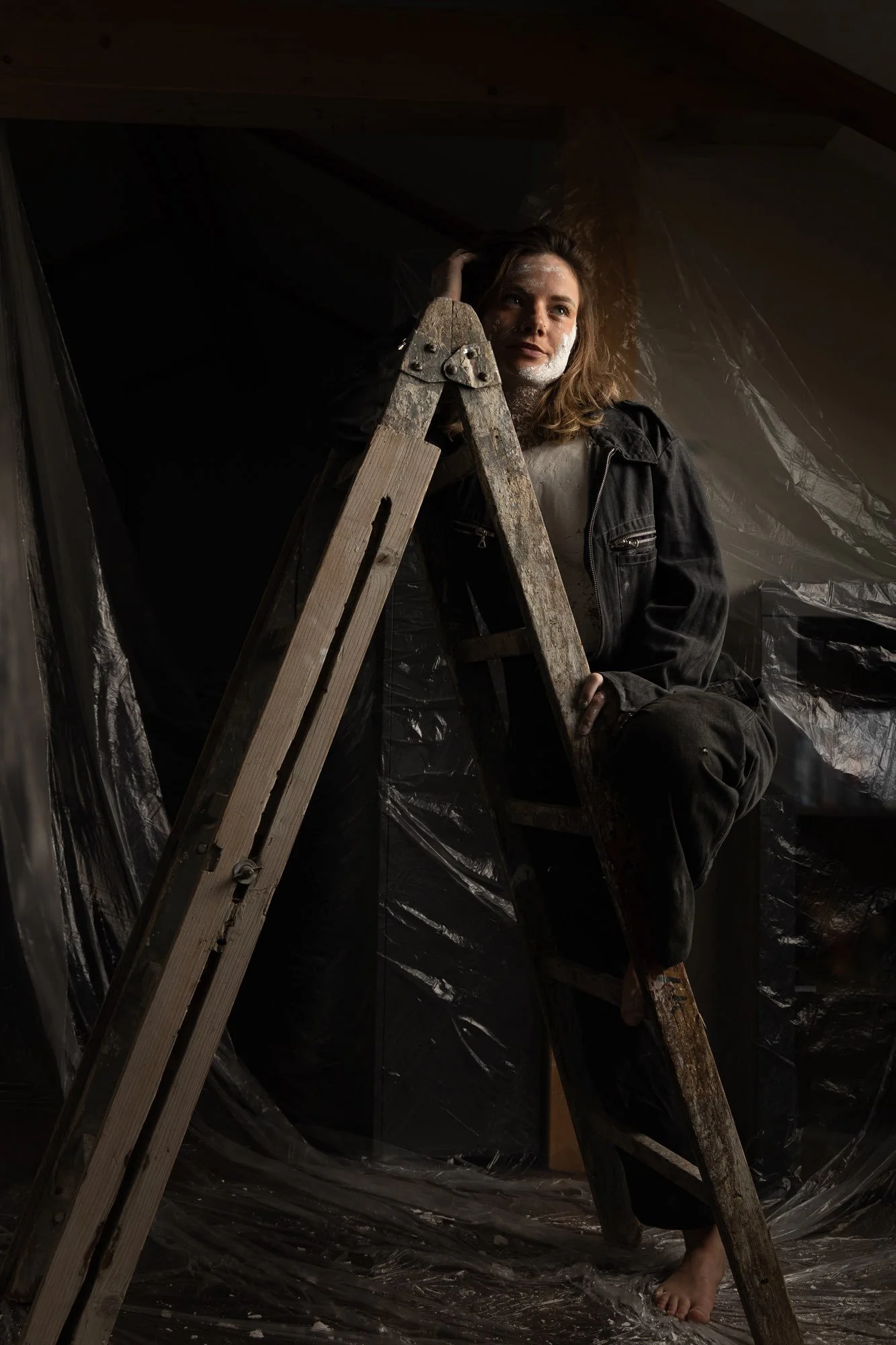 Junge Frau sitzt auf einer Leiter, trägt eine schwarze Lederjacke und eine weiße Bluse. Die Szene ist im Dunkeln mit Plastikfolie im Hintergrund, und sie hat weißen Puder im Gesicht.