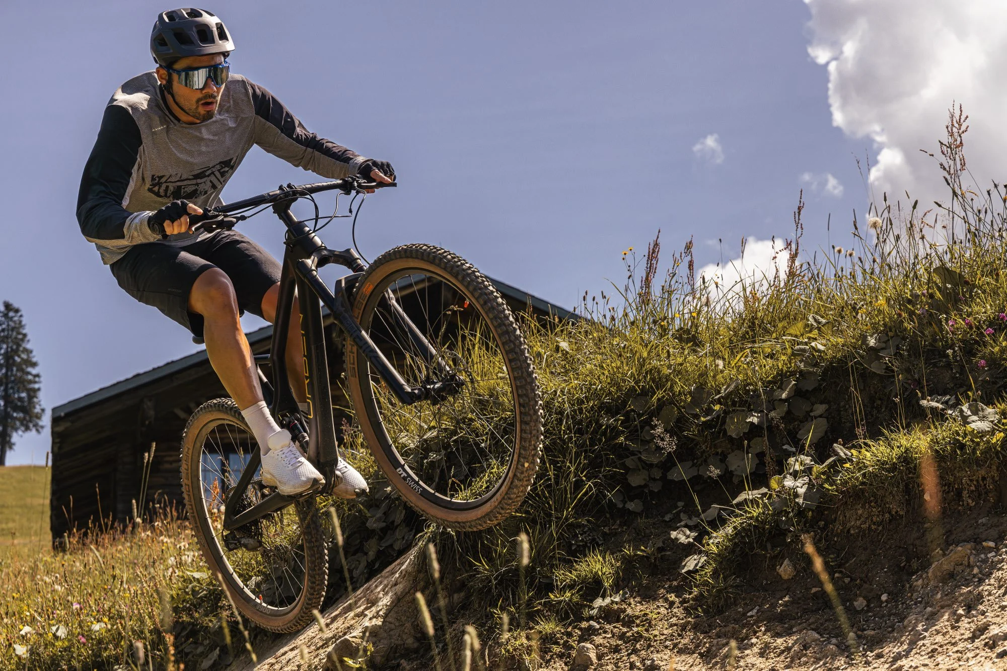 Mann fährt Mountainbike auf steilem Hang, trägt Helm und Schutzbrille, im Hintergrund ein Haus und blauer Himmel